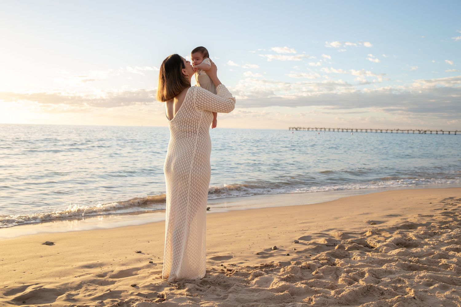 Woman in white dress holds baby up on sandy beach at golden sunset with pier in background.