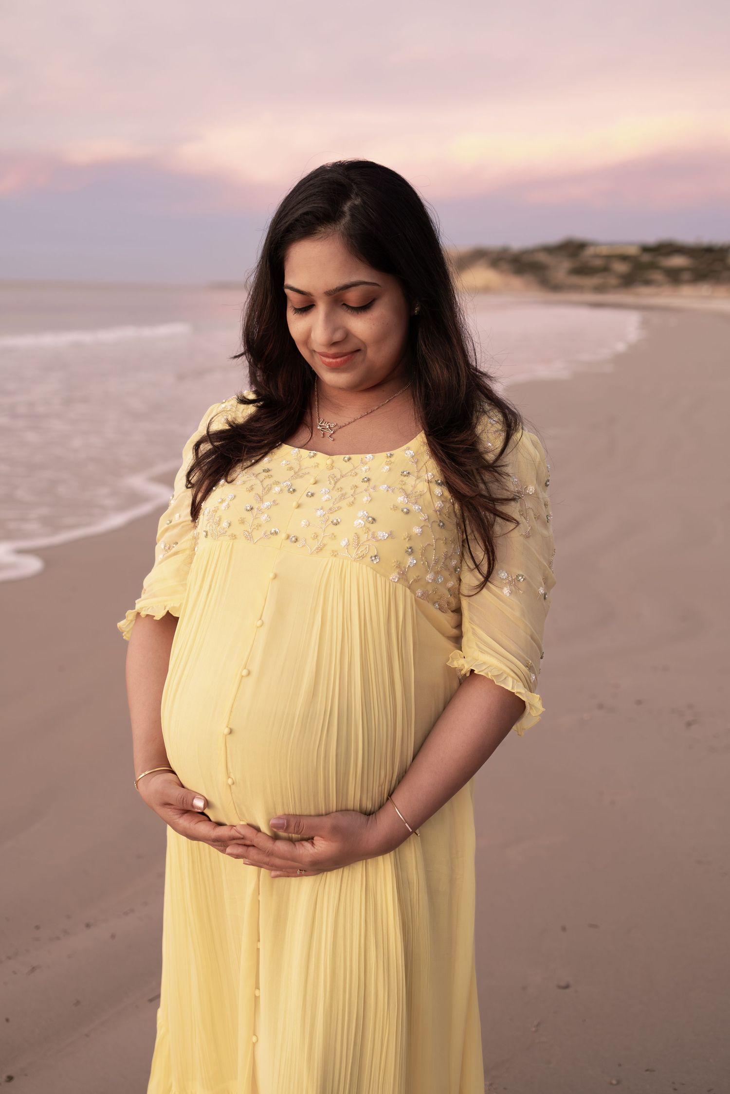 Pregnant woman in yellow dress holding belly on beach at sunset.