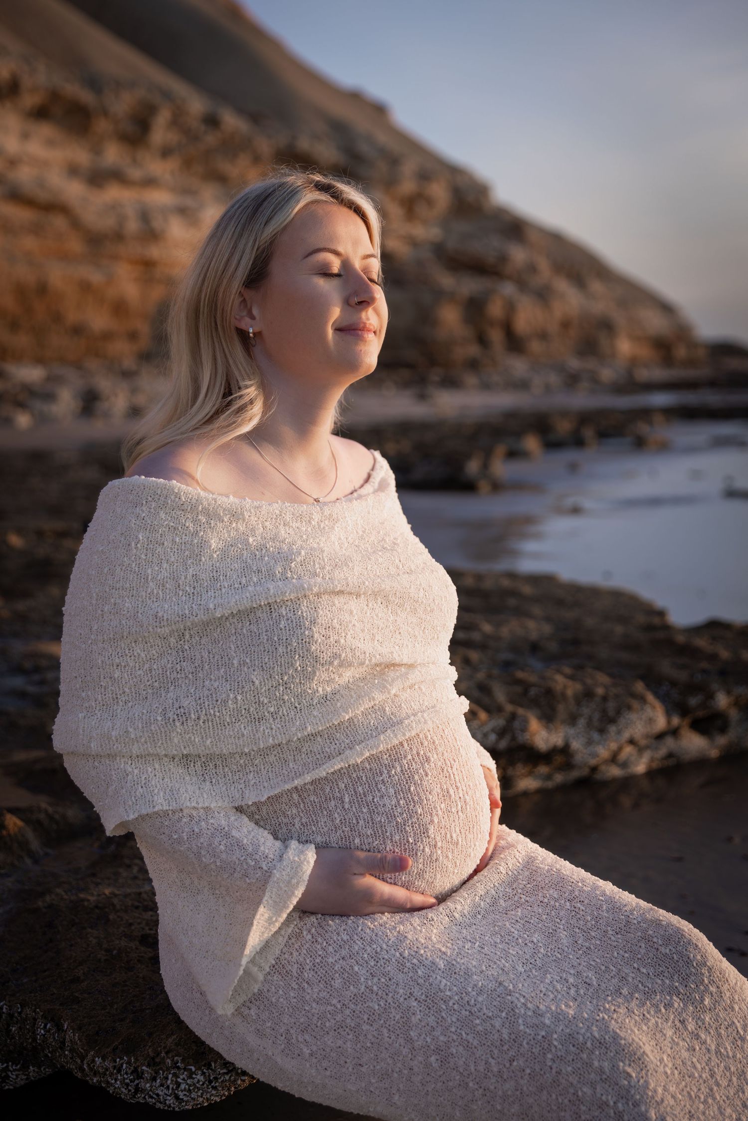 Maternity photo session at sunset featuring a stretchy knit white dress against rocky coastal cliffs.