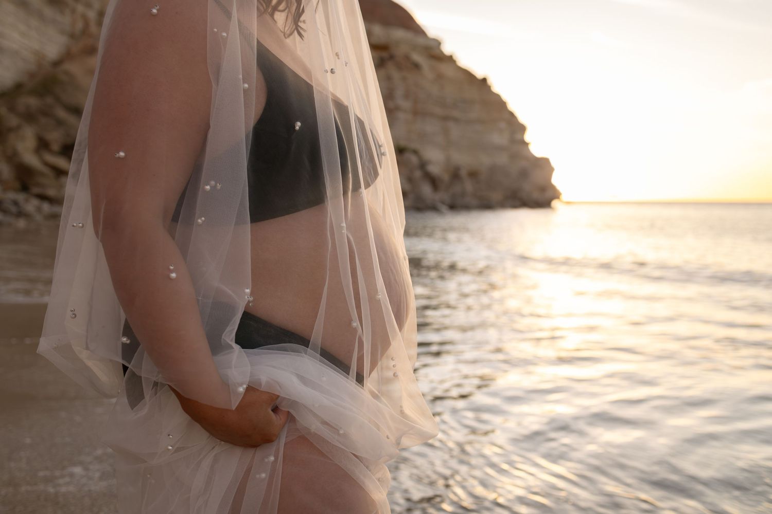 A pregnant mother in flowy veil stands on a beach at sunset with rocky cliffs in the background.