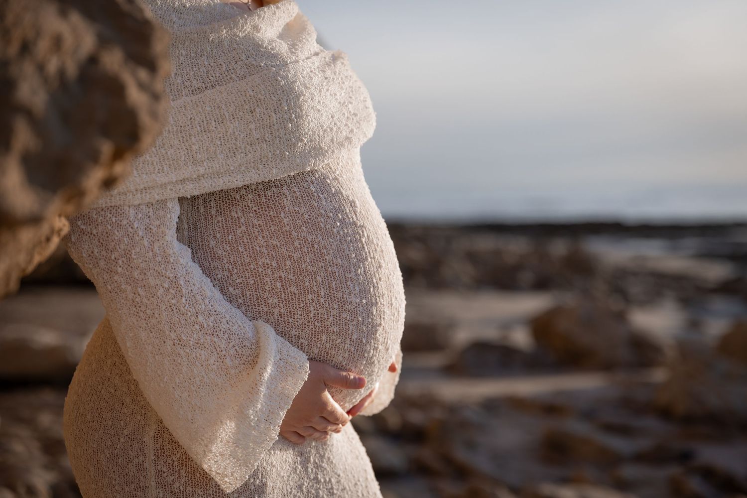 A pregnant woman in a white dress stands on a rocky beach at sunset with waves crashing in the background.