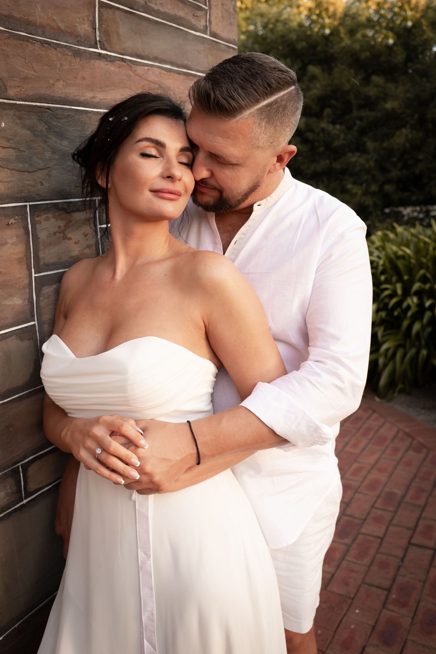 Romantic couple in white outfits embracing against brick wall during sunset photoshoot.