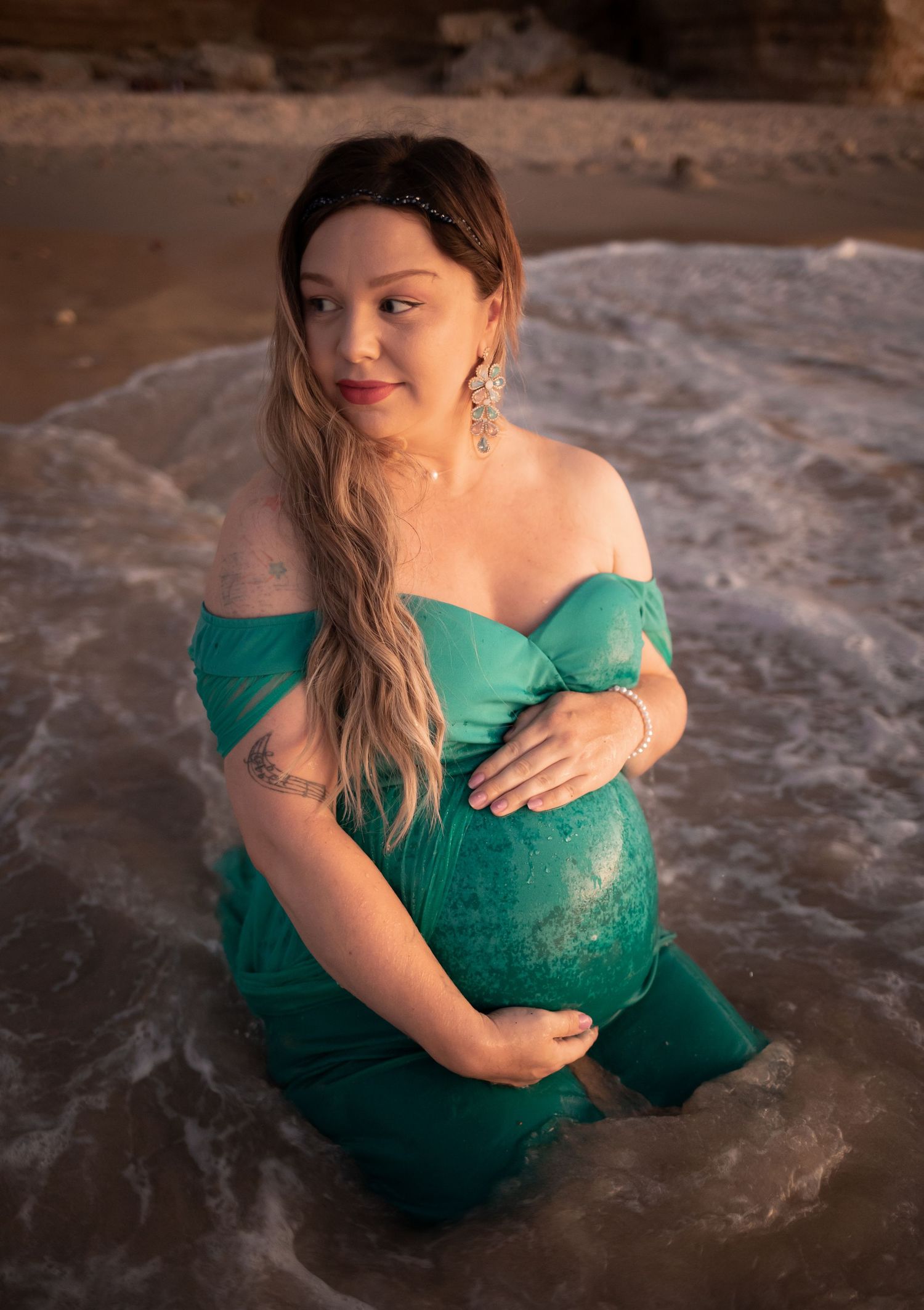 Pregnant woman in emerald dress poses in shallow ocean water during sunset.
