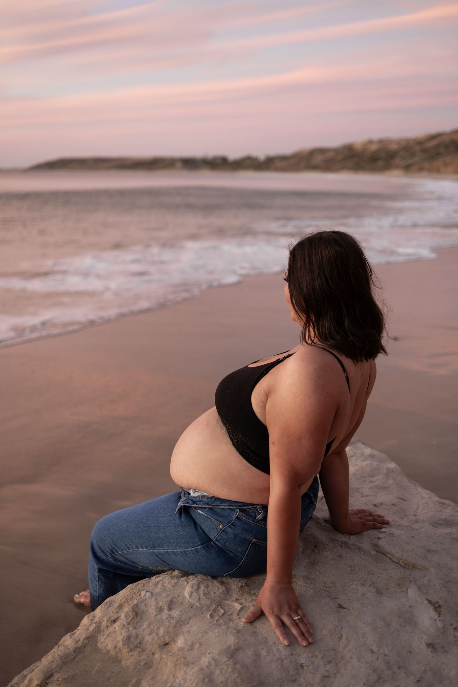 A pregnant woman sits on coastal rocks during sunset, gazing at the ocean.