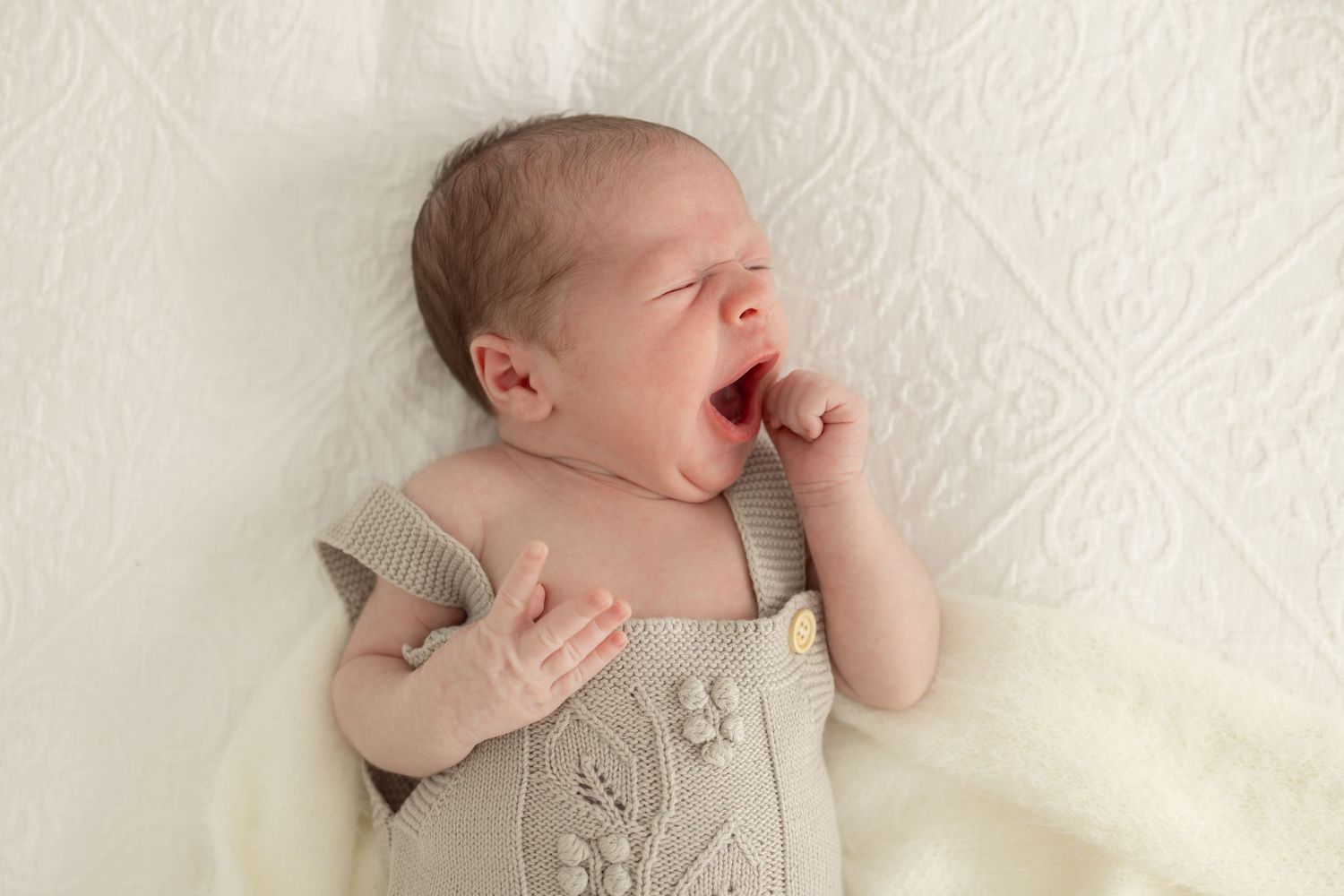 Baby in beige knit romper outfit lying on textured white background.