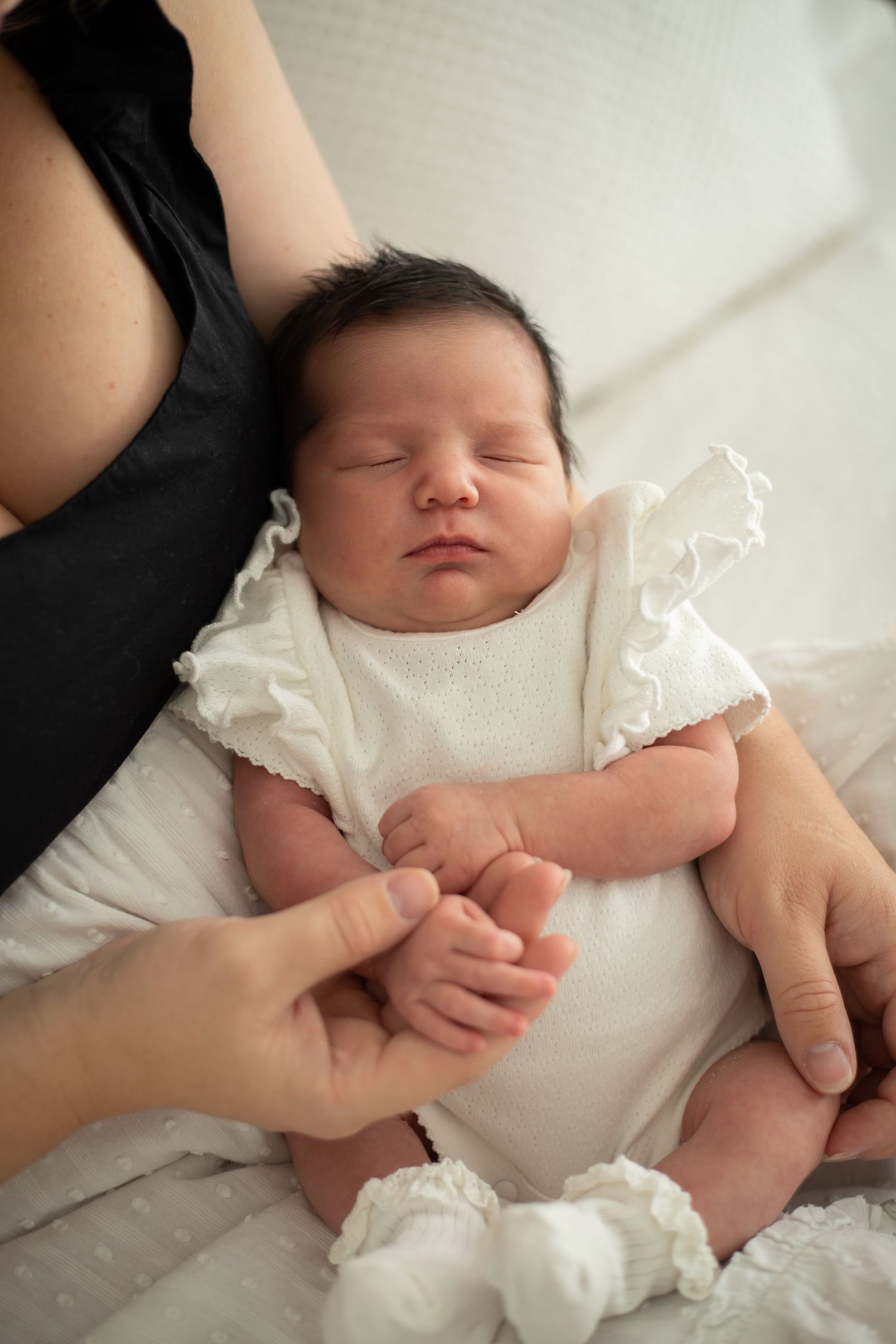 Newborn baby wearing white outfit resting peacefully in mothers lap on white bedding.