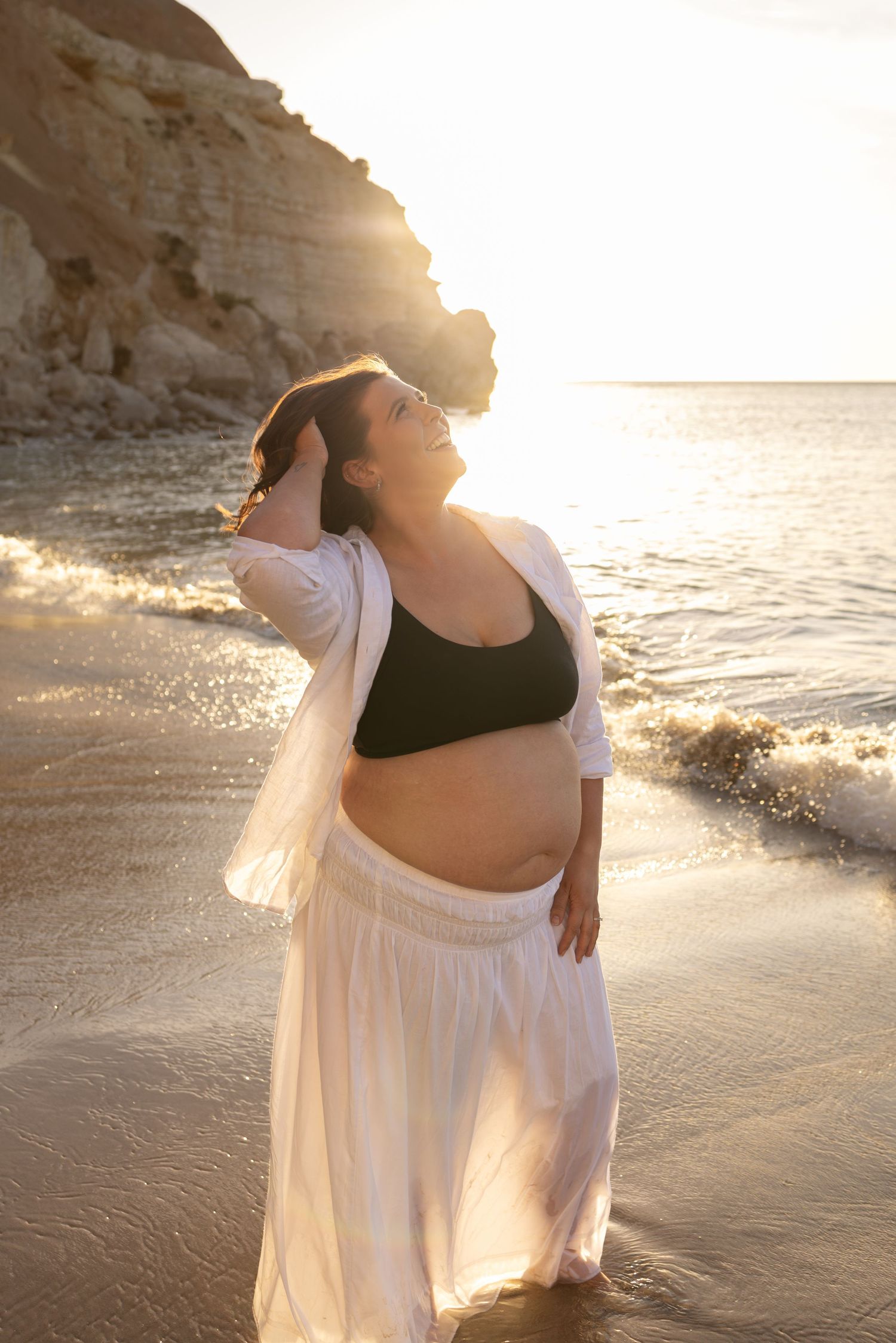Sunset maternity beach photos with flowing white skirt and black crop top against ocean waves.
