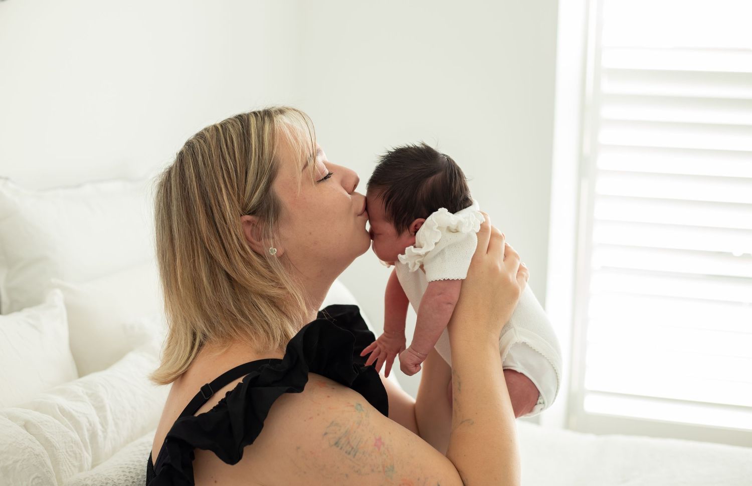 Tender moments of mother kissing newborn baby in bright white bedroom setting.