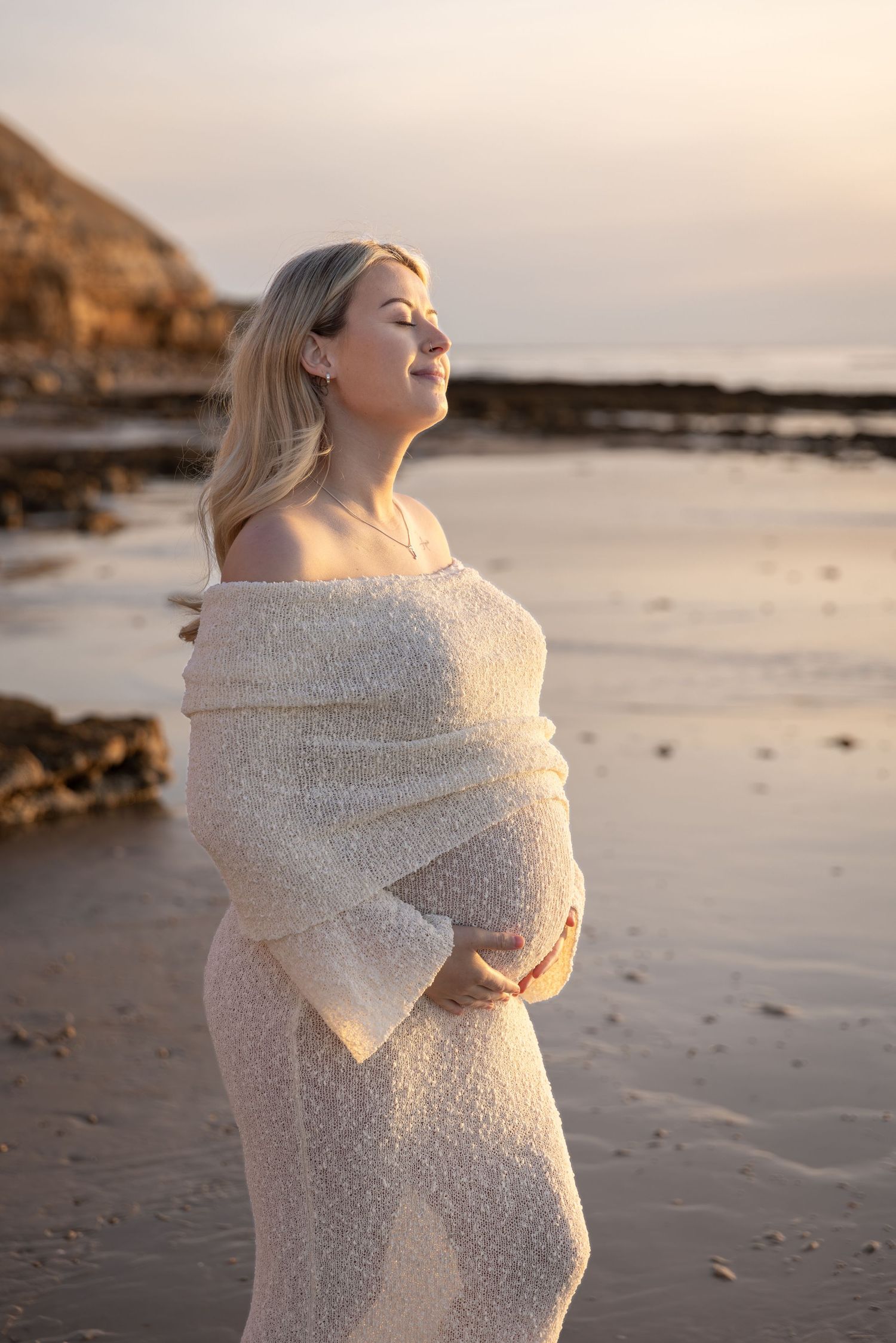 Pregnant woman in white wrap standing on beach at sunset, cradling her belly with eyes closed peacefully.
