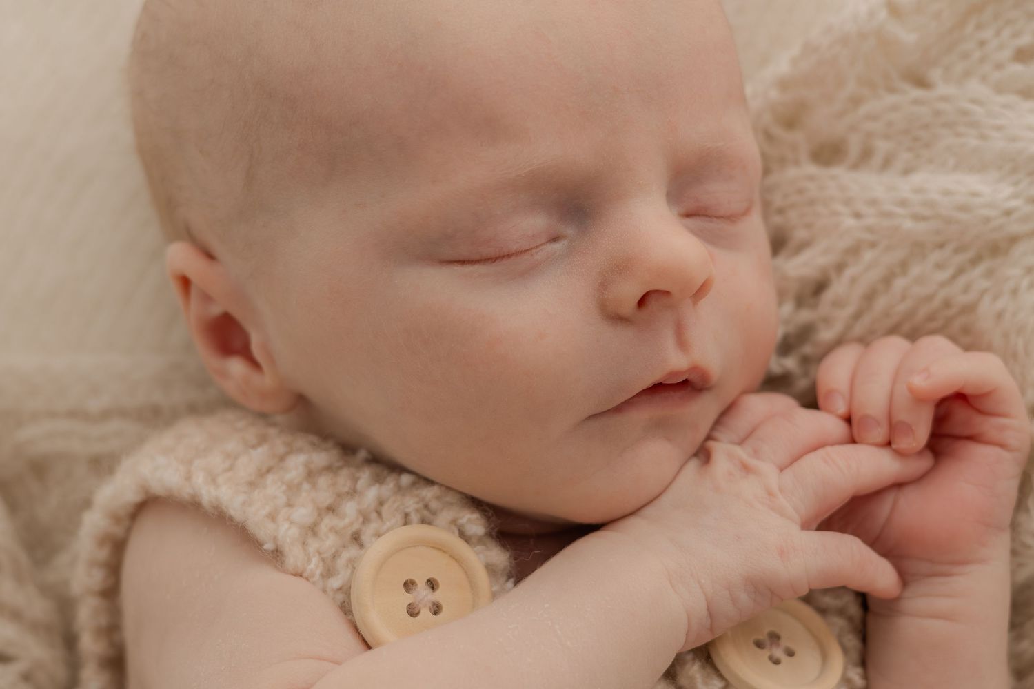 Sleeping newborn wearing knit romper outfit with wooden button detail on lace blanket.