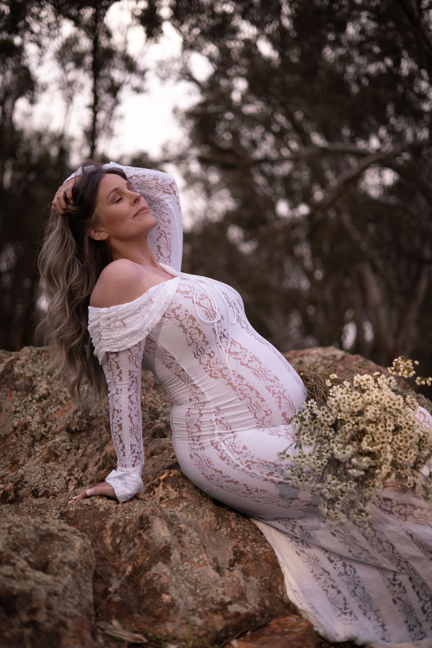 Ethereal maternity portrait featuring a woman with hands in hair wearing white lace dress reclining on rocky terrain.