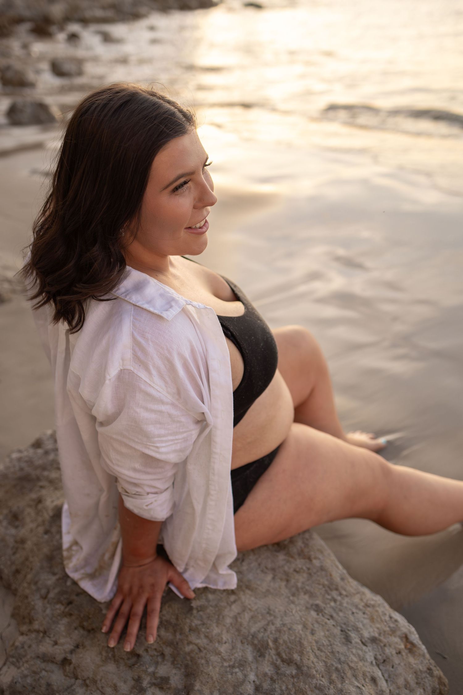 Candid maternity photos on rocky beach during golden hour wearing white shirt and black underwear.