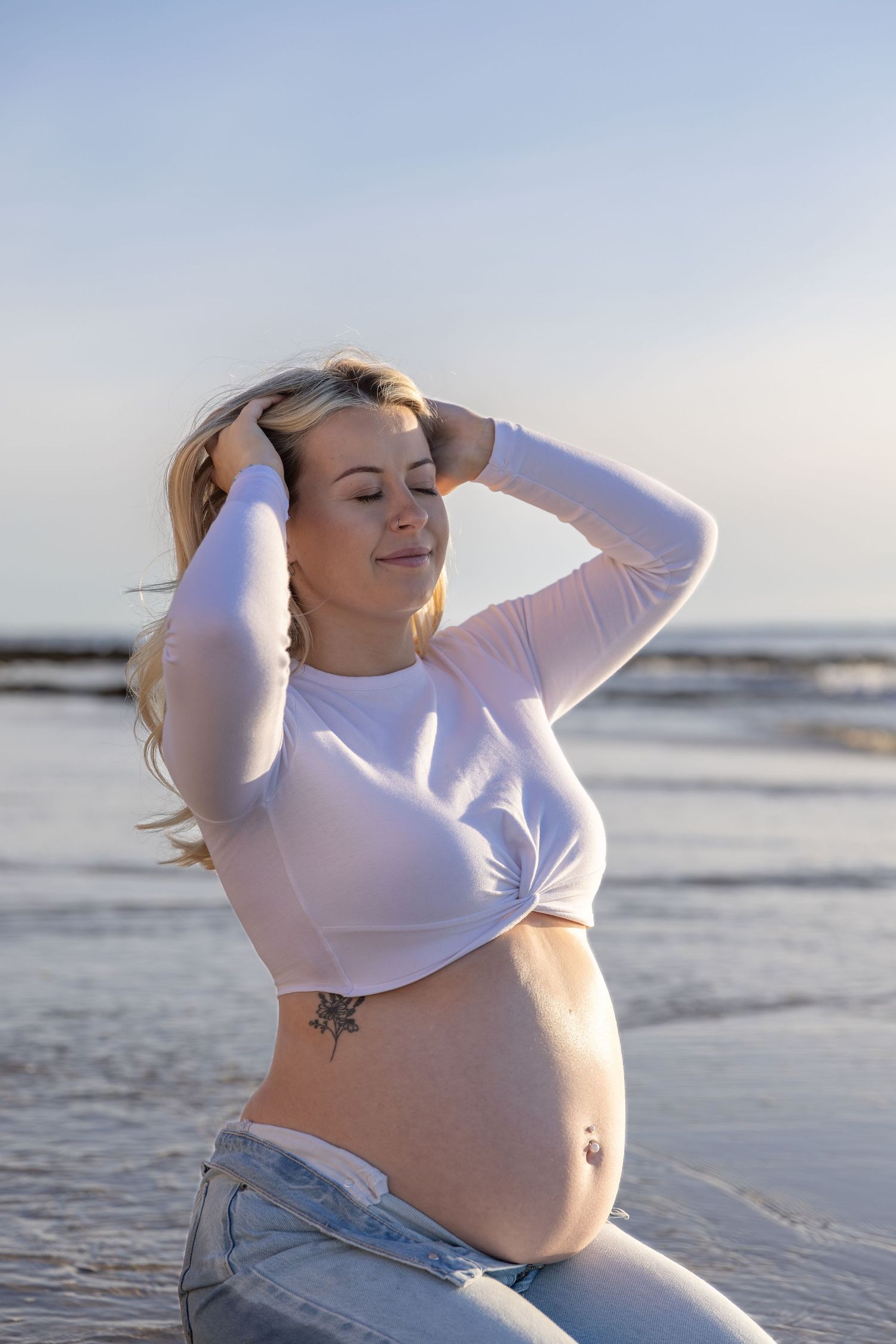 Beachside maternity portrait of mother holding hair in casual white top and denim.