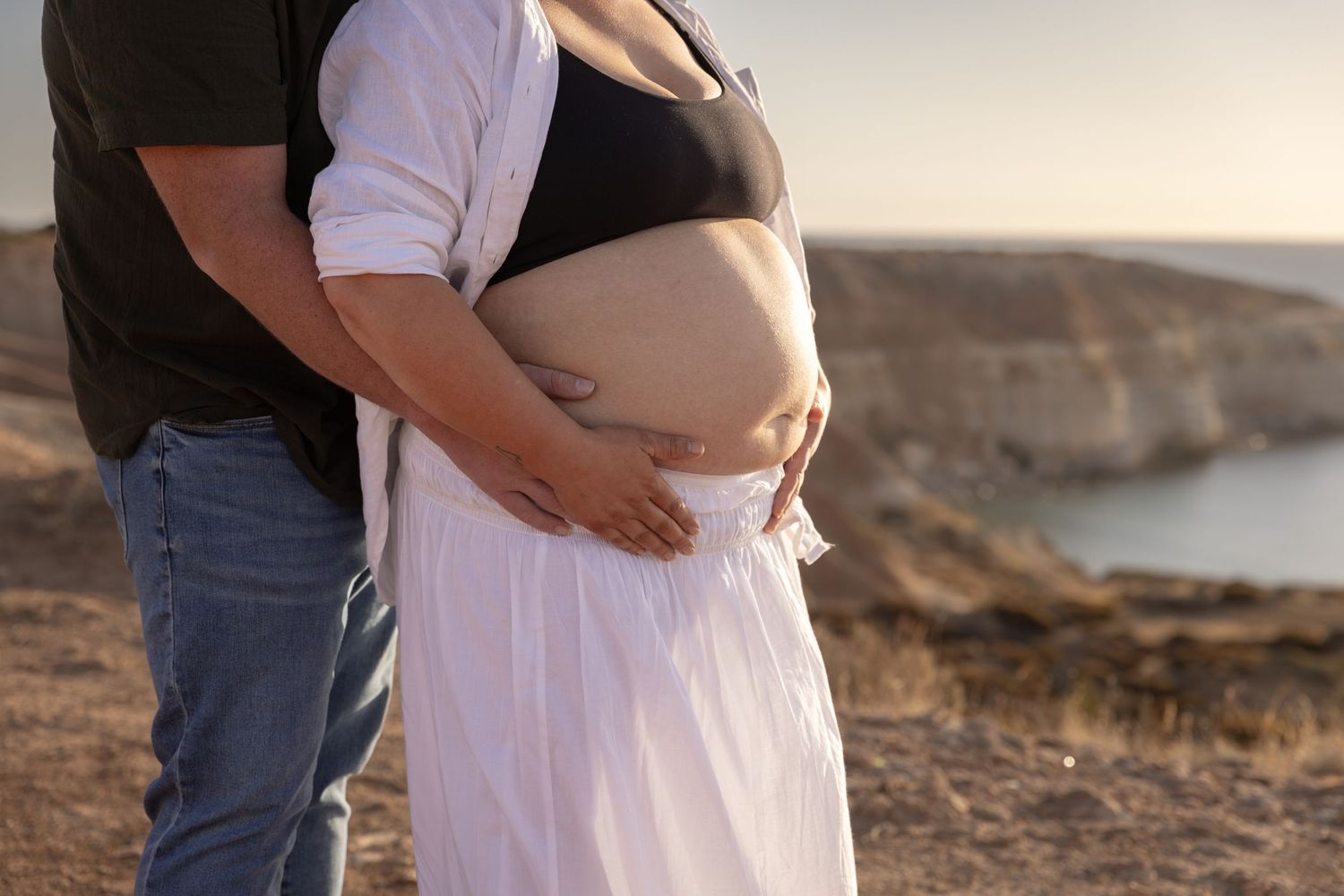 Maternity photoshoot at a rocky beach during sunset with couple embracing and wearing casual attire.