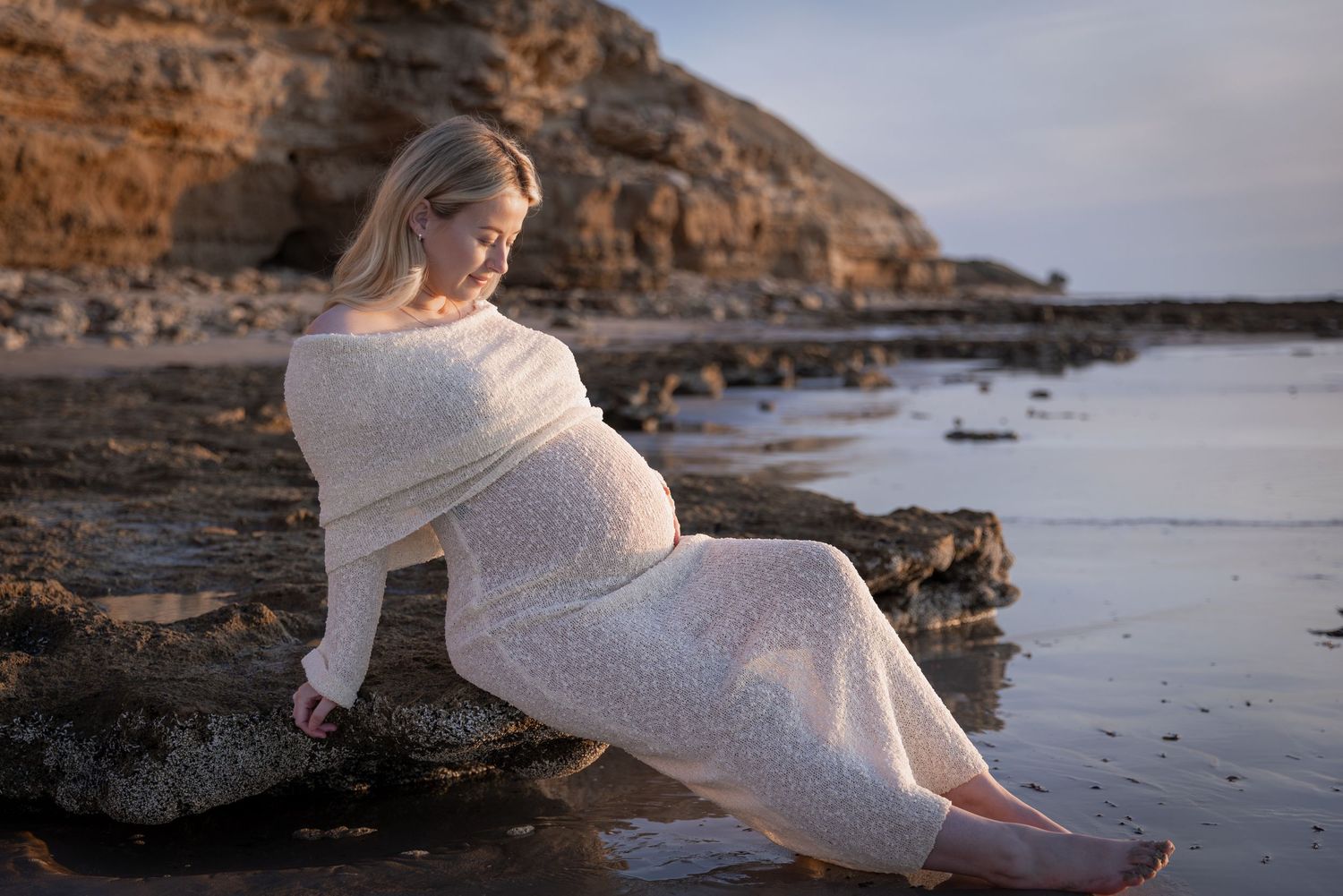 Artistic maternity photos on rocky coastline with woman in ethereal white knit dress.