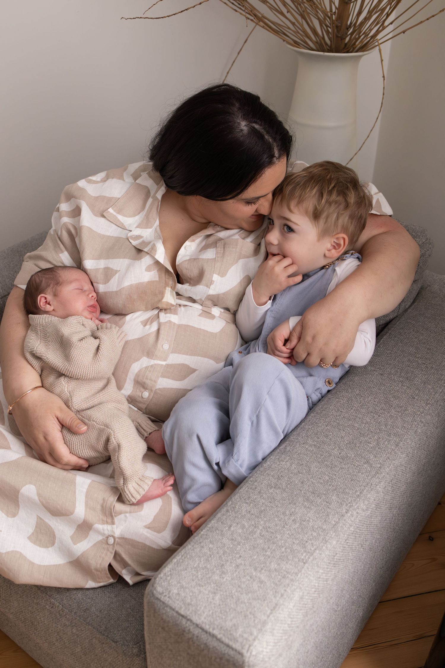 A tender family moment on a gray armchair with a newborn and toddler.