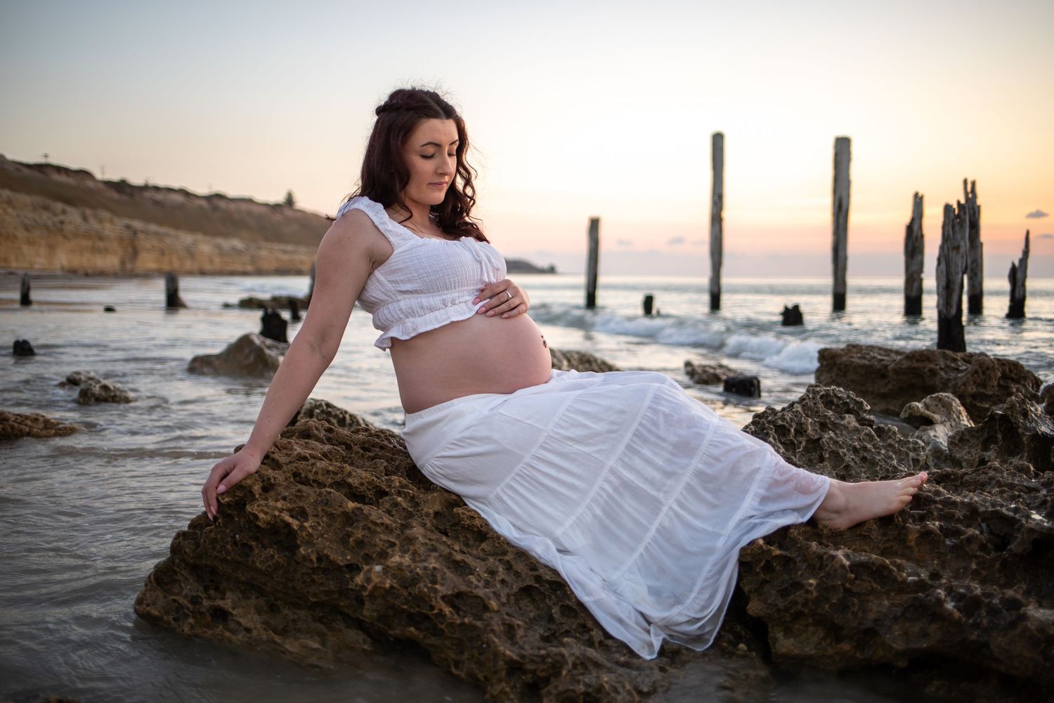 Artistic maternity photos on beach with weathered wooden posts at sunset.