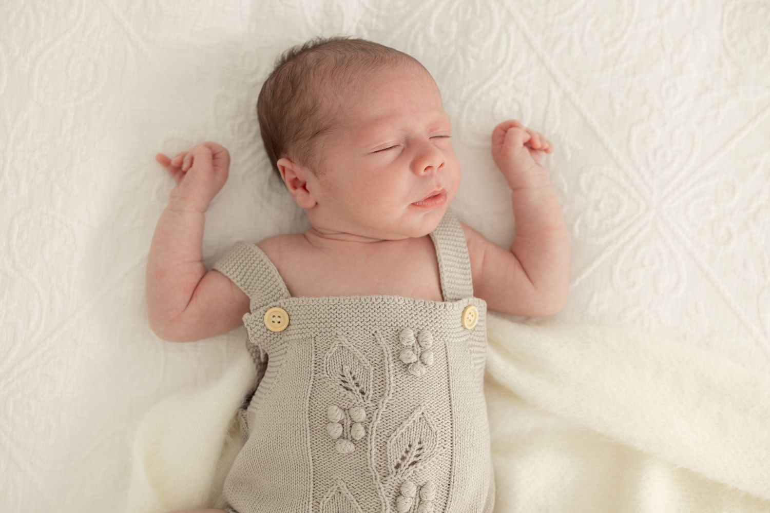 Newborn baby resting on white textured blanket wearing beige knitted romper outfit.