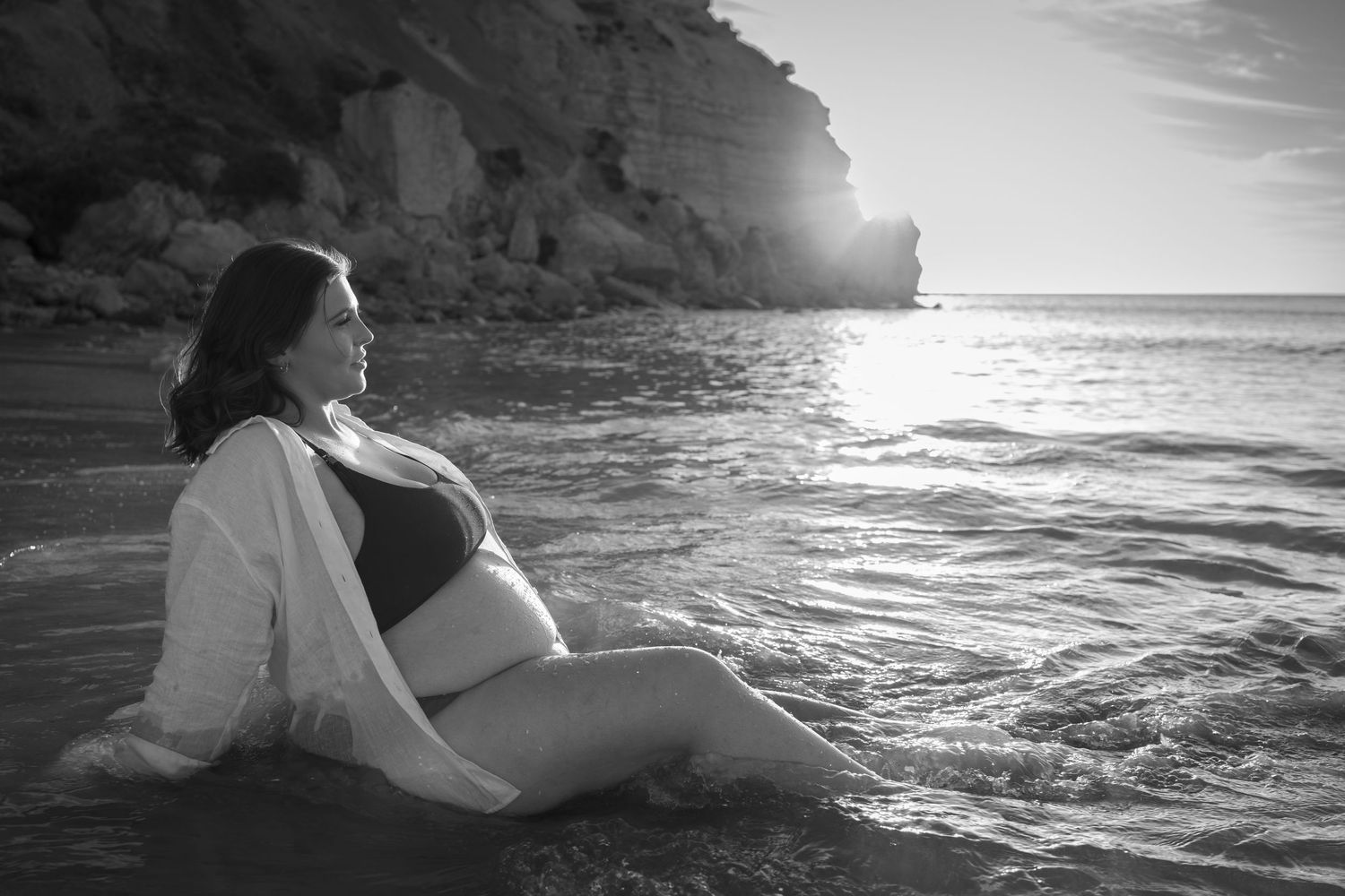 Black and white scenic beach maternity session with rocky cliffs and ocean waves at sunset.