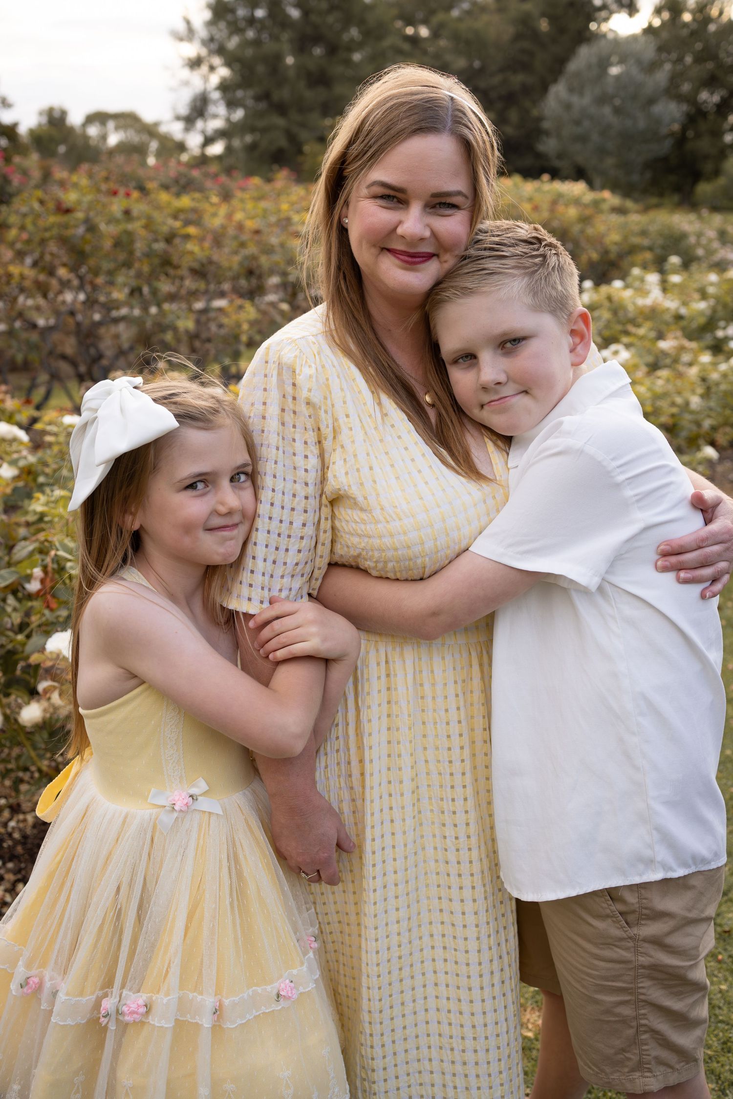 A woman in a yellow dress smiles with two children hugging her in a lush garden setting.