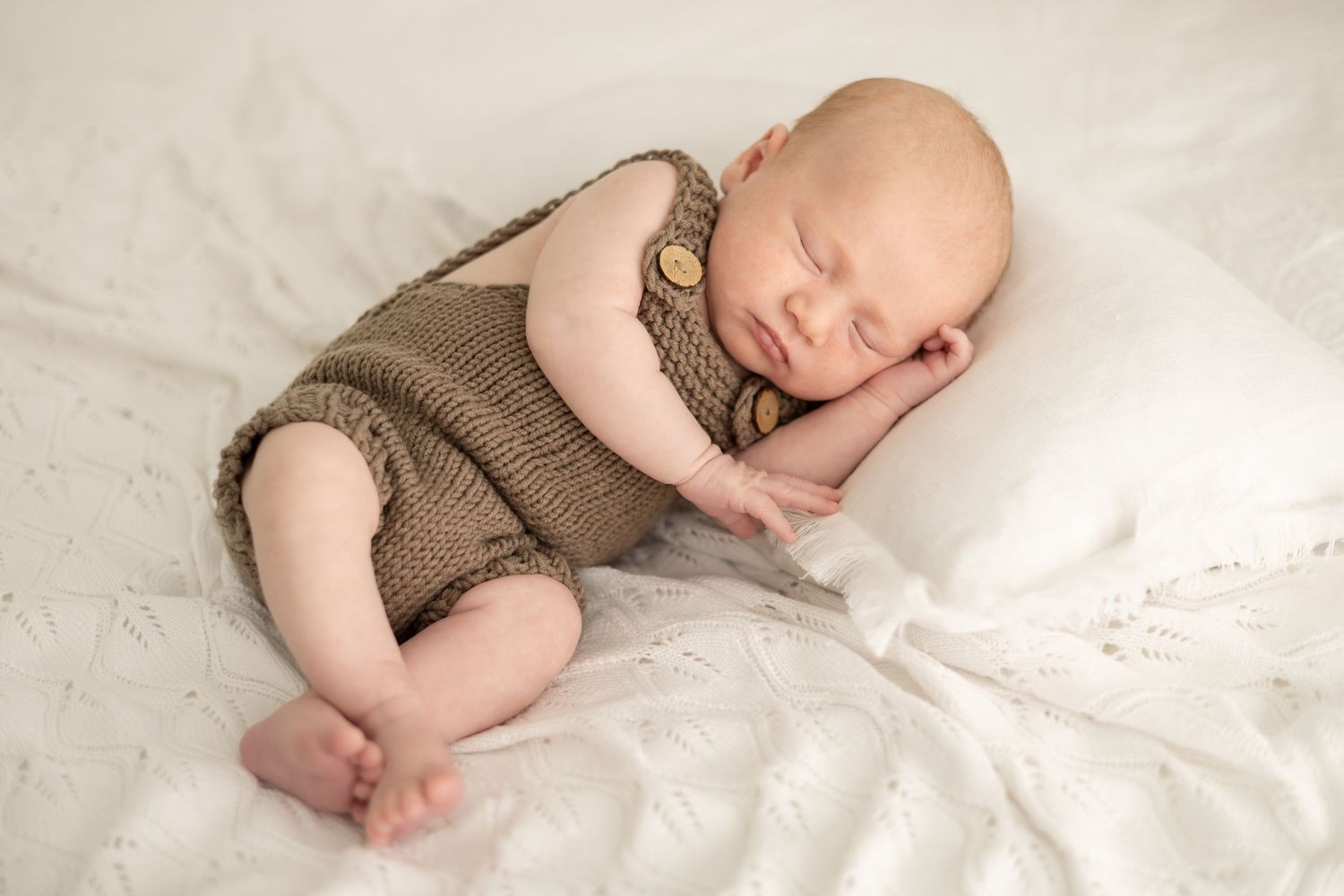 Newborn baby sleeping peacefully on white blanket wearing brown knit romper outfit.