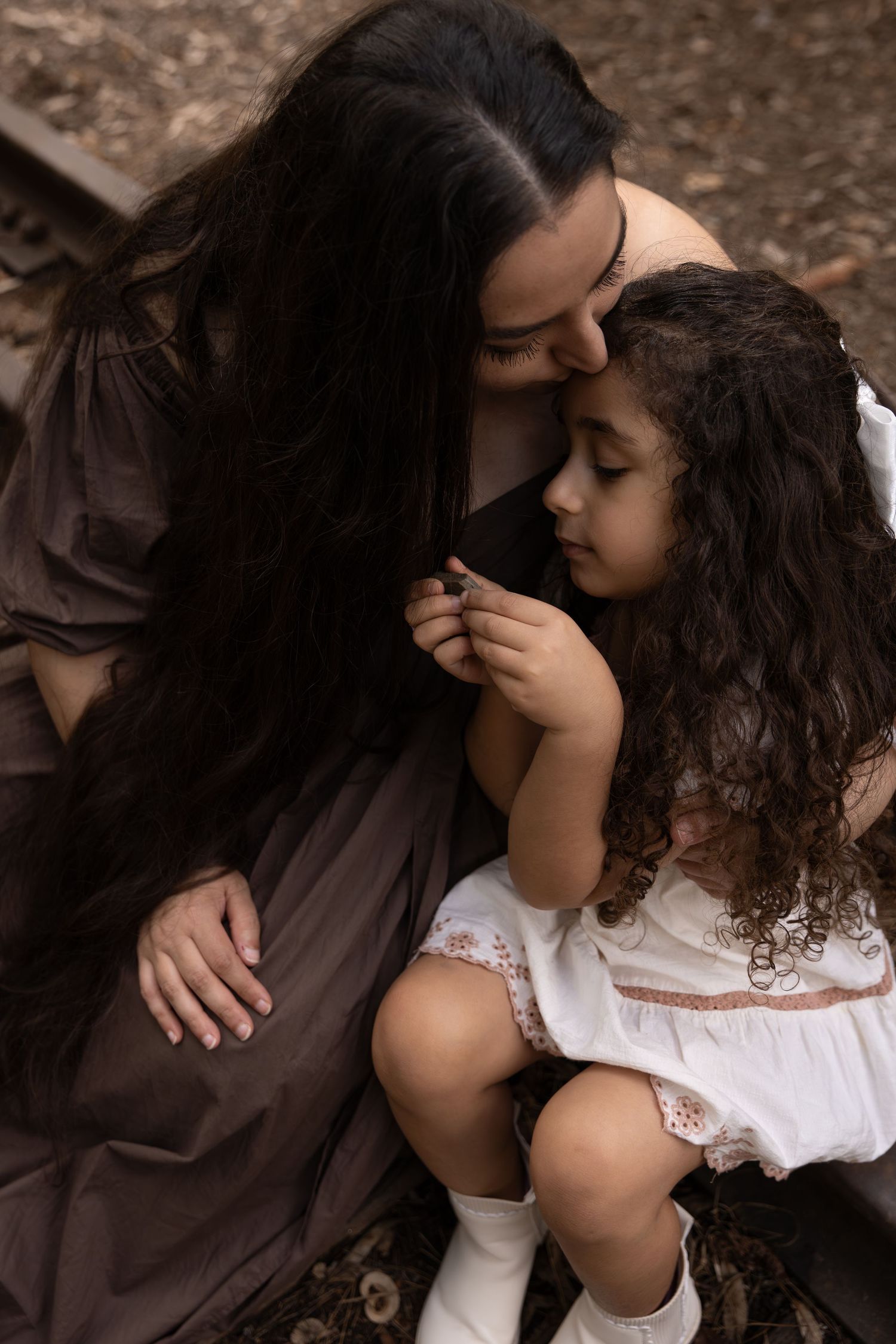 A mother and young daughter with curly hair share a tender moment outdoors, dressed in brown and white dresses.