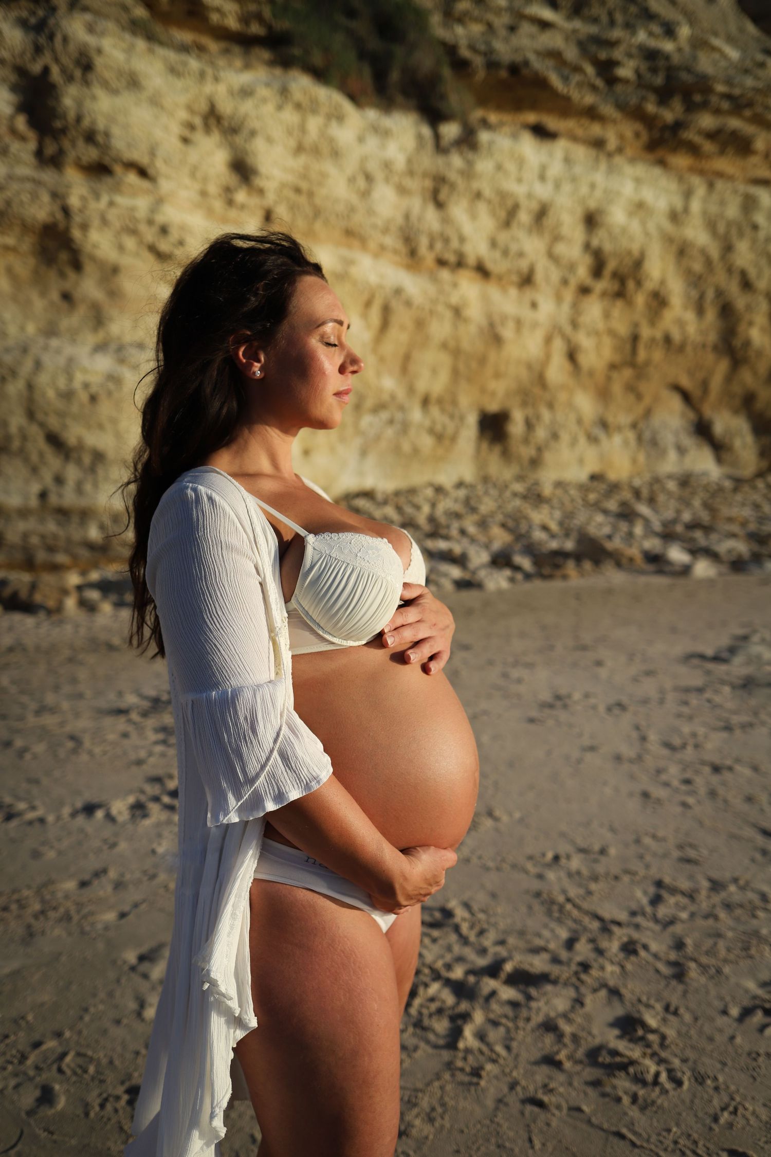 Maternity portrait at beach with white flowing robe and white underwear against rocky cliff background.