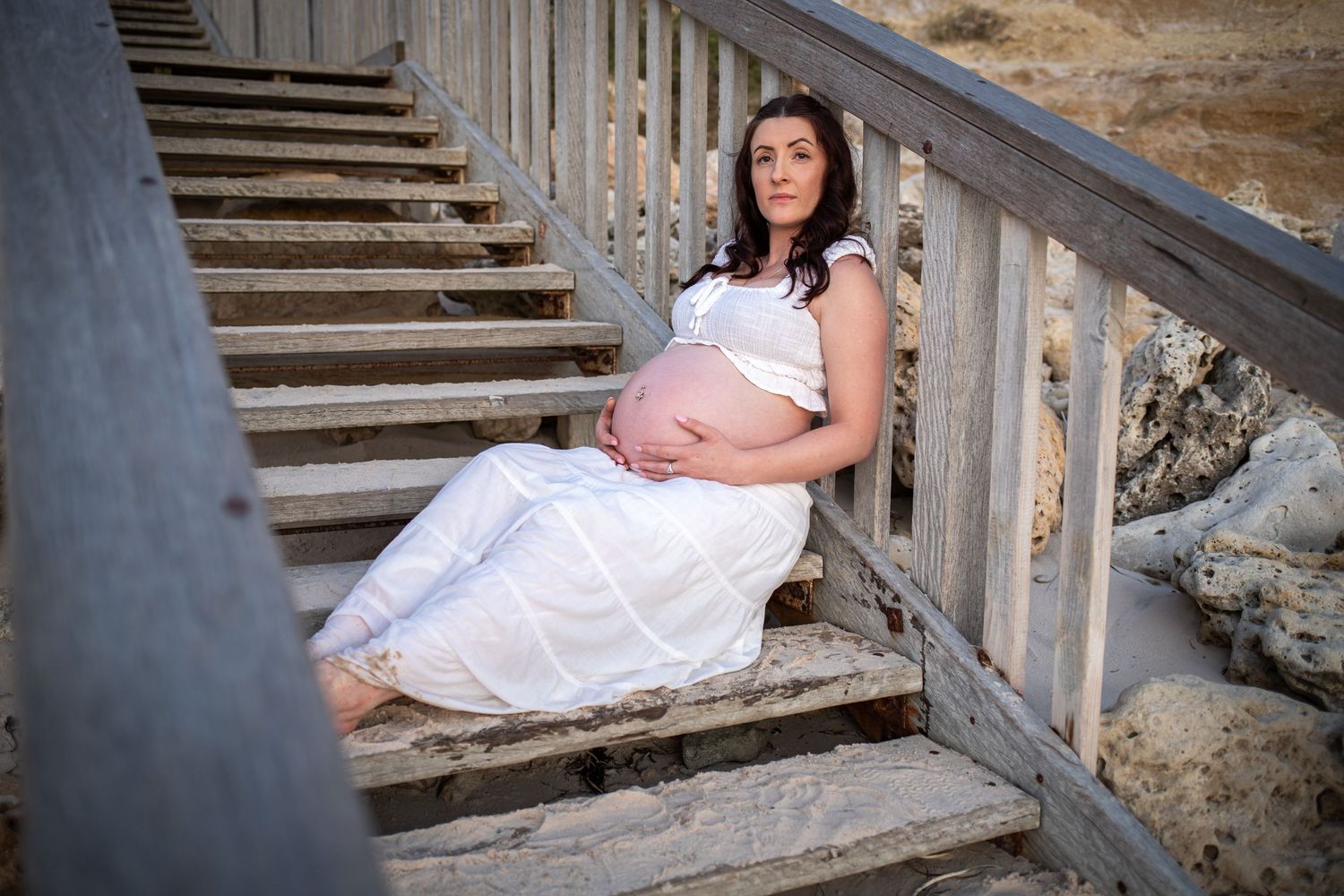 Maternity photoshoot on weathered wooden beach stairs in soft evening light.