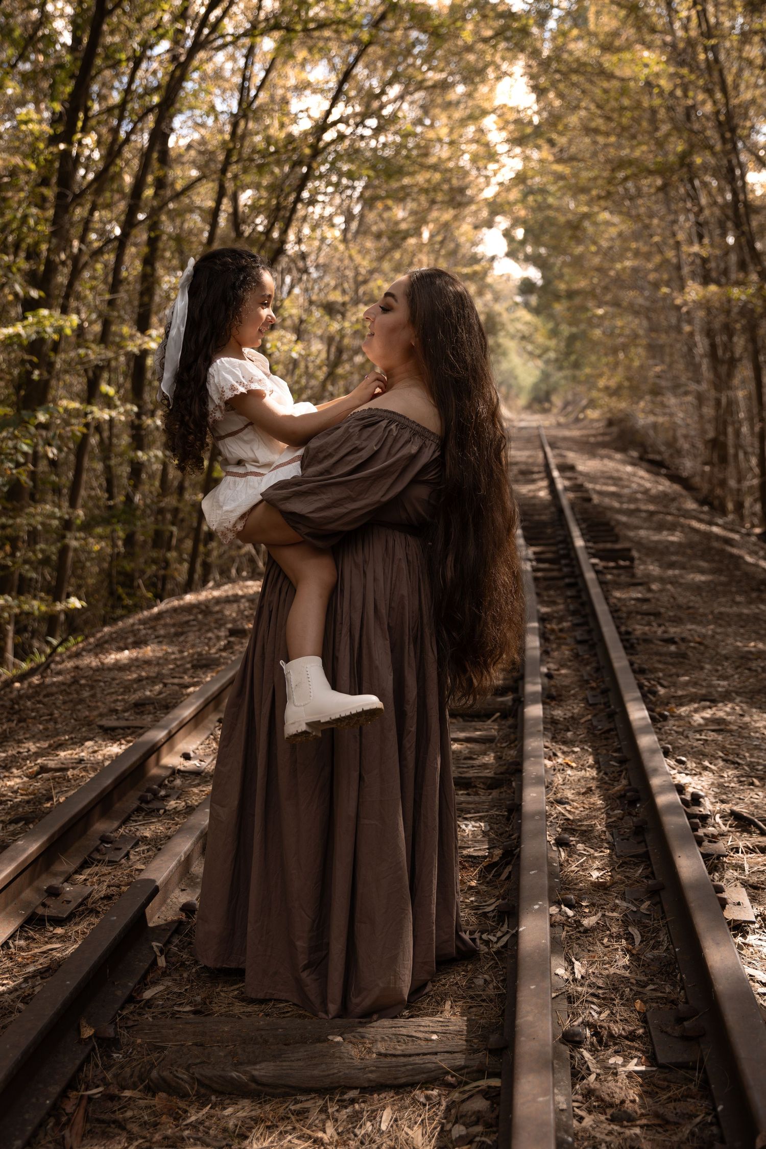 A woman in a brown gown holds a young girl on railroad tracks surrounded by autumn trees.