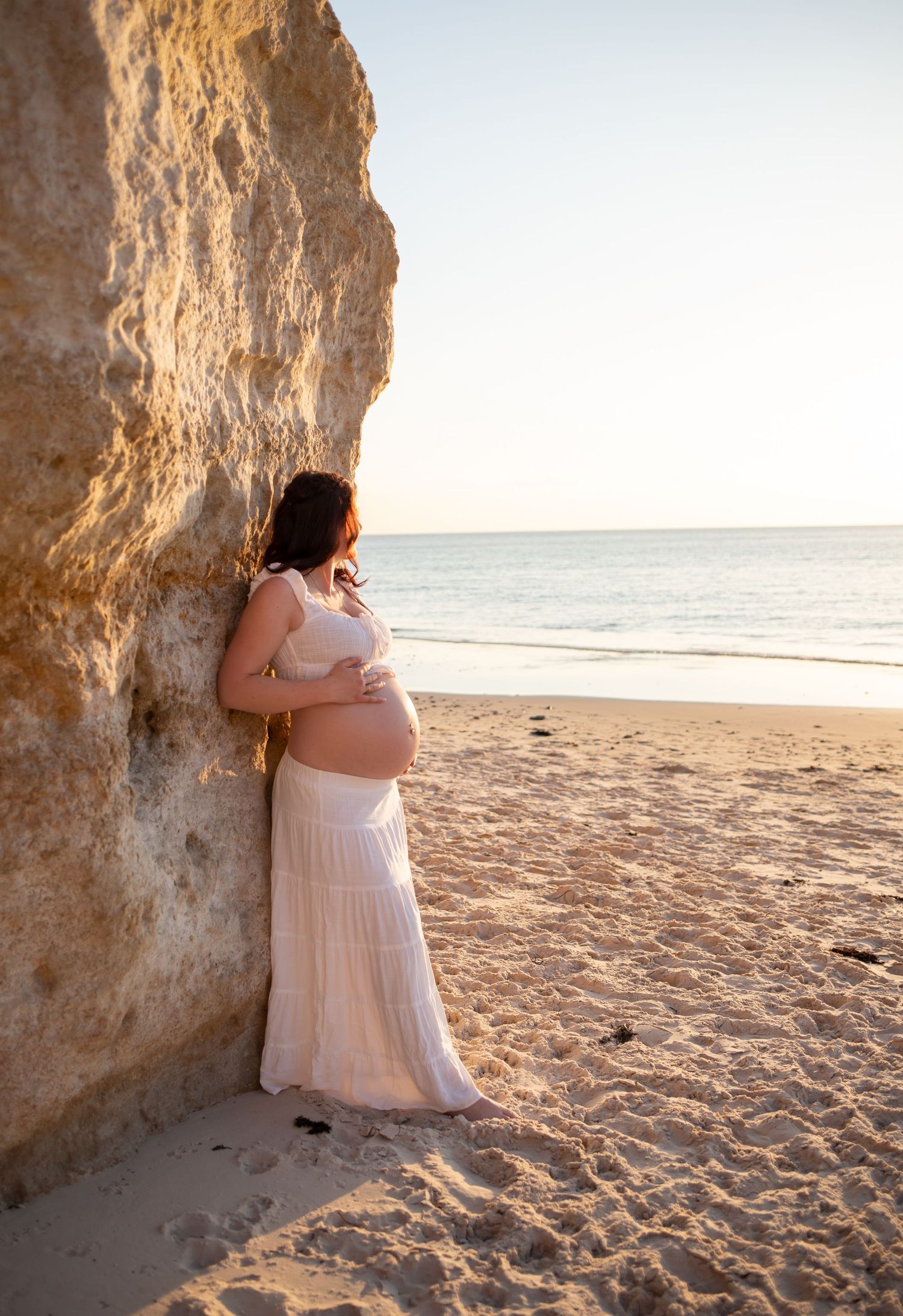 Expectant mother in flowing white skirt poses against rocky beach cliffs at sunset.