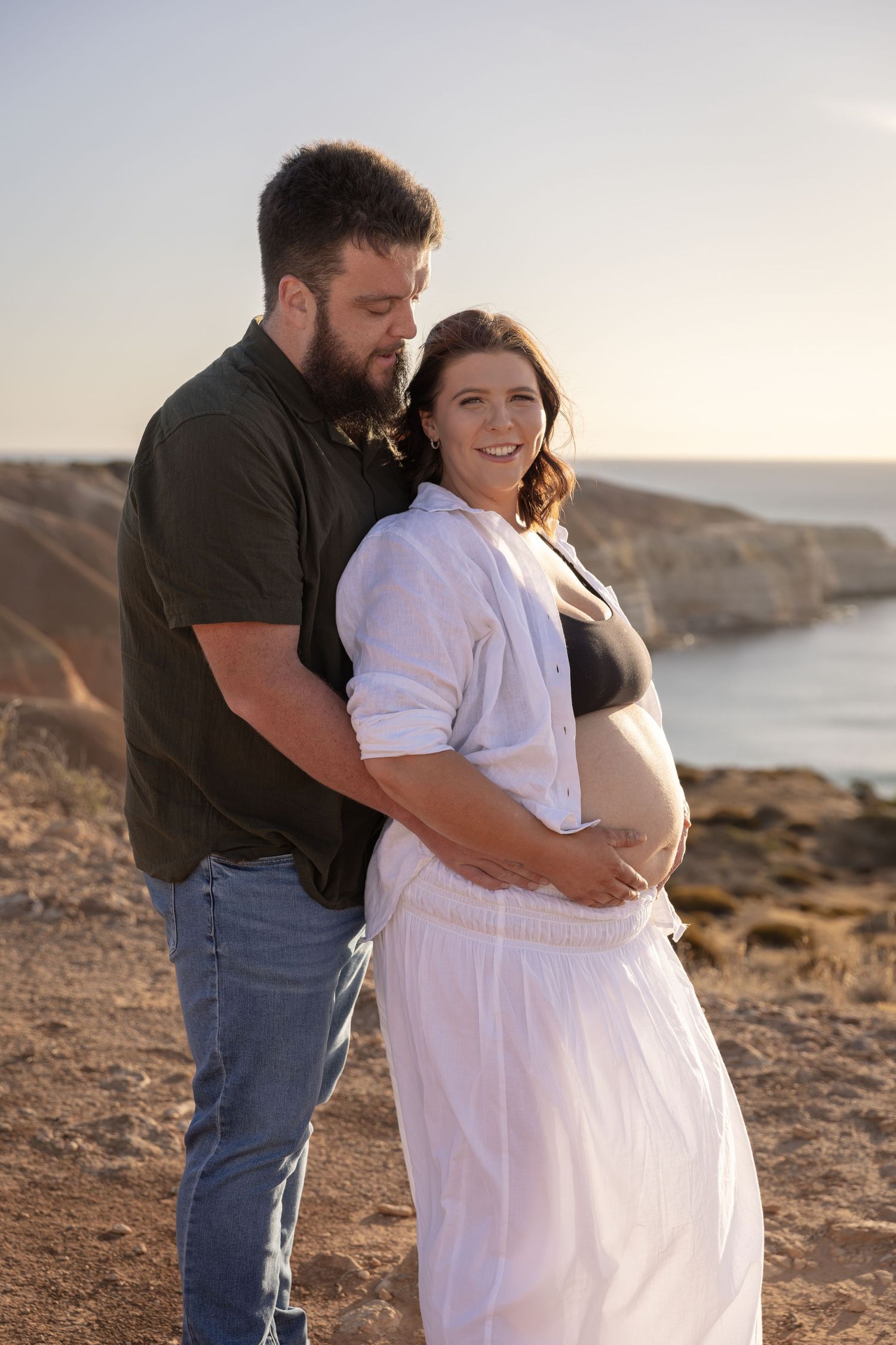 Couple embracing during sunset maternity photoshoot on rocky coastline.