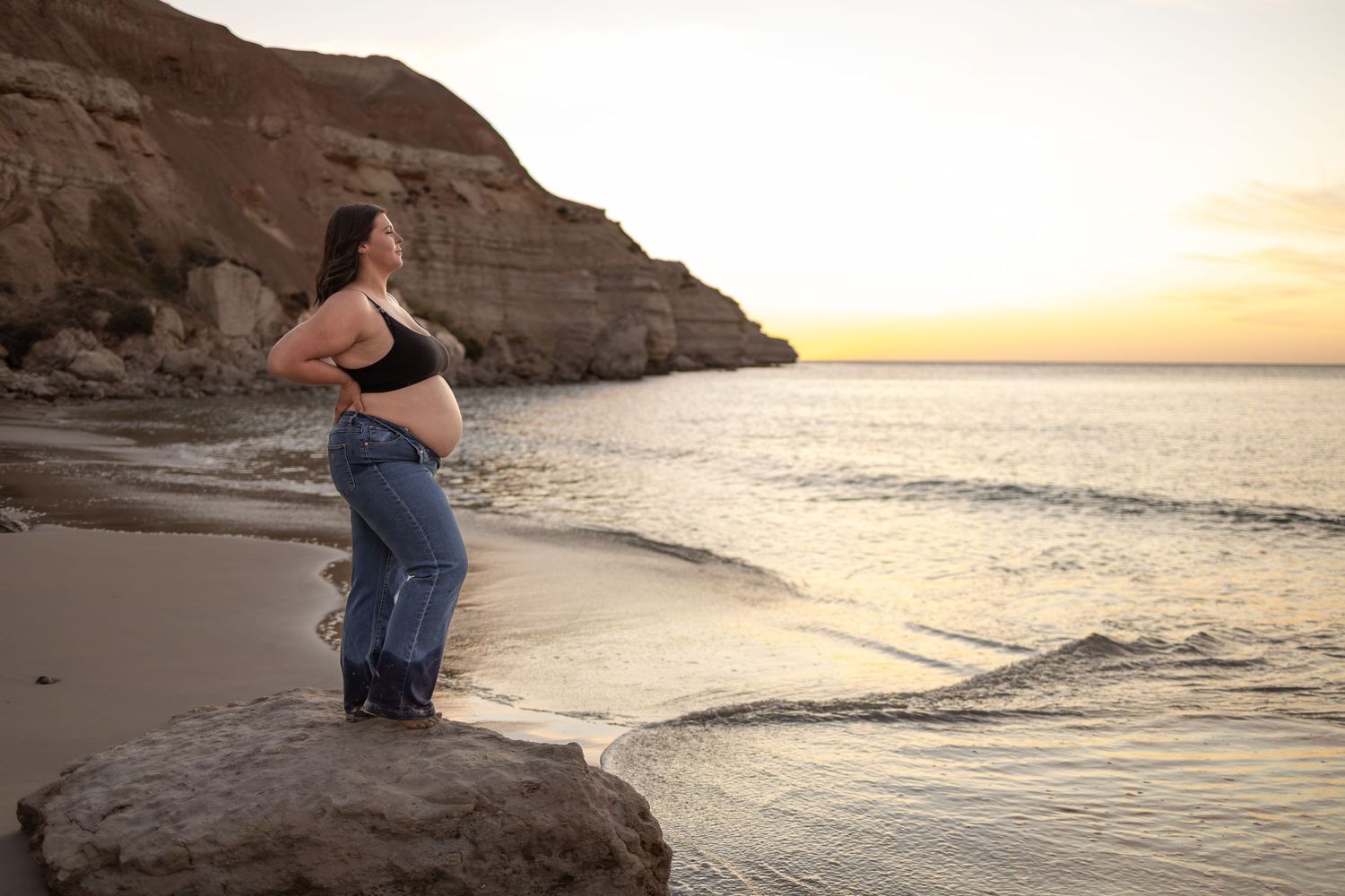 Silhouette stands on rocky outcrop overlooking ocean at sunset during maternity photoshoot.