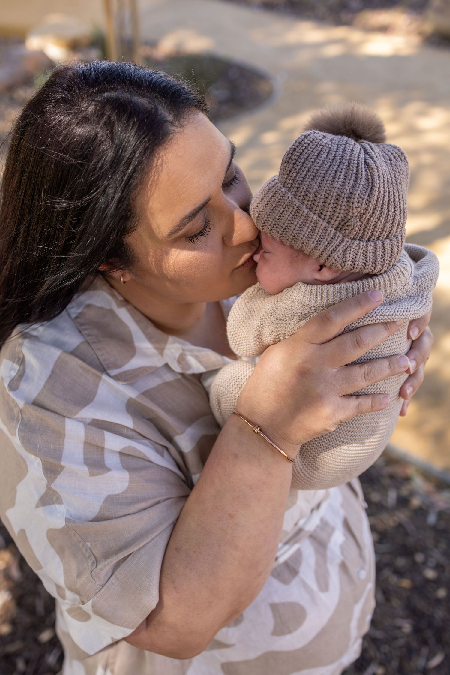 Mother in patterned dress kisses baby wearing knitted beanie outdoors.