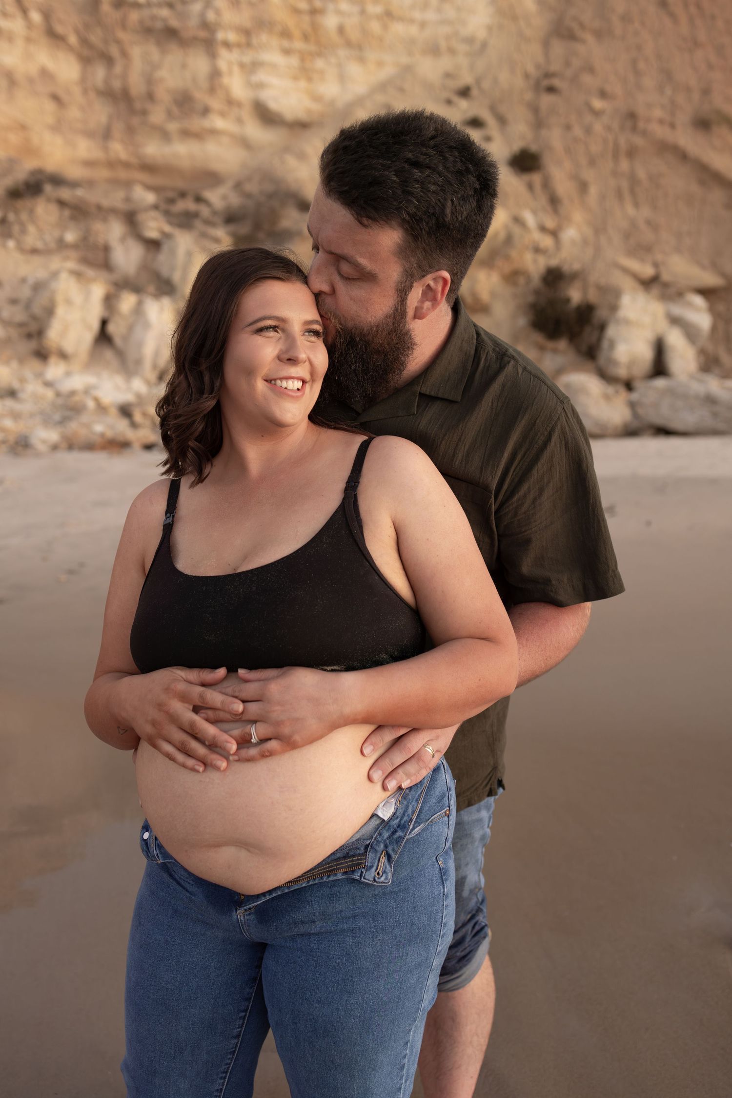 Couple embraces during sunset maternity photoshoot on sandy beach with cliffs behind.