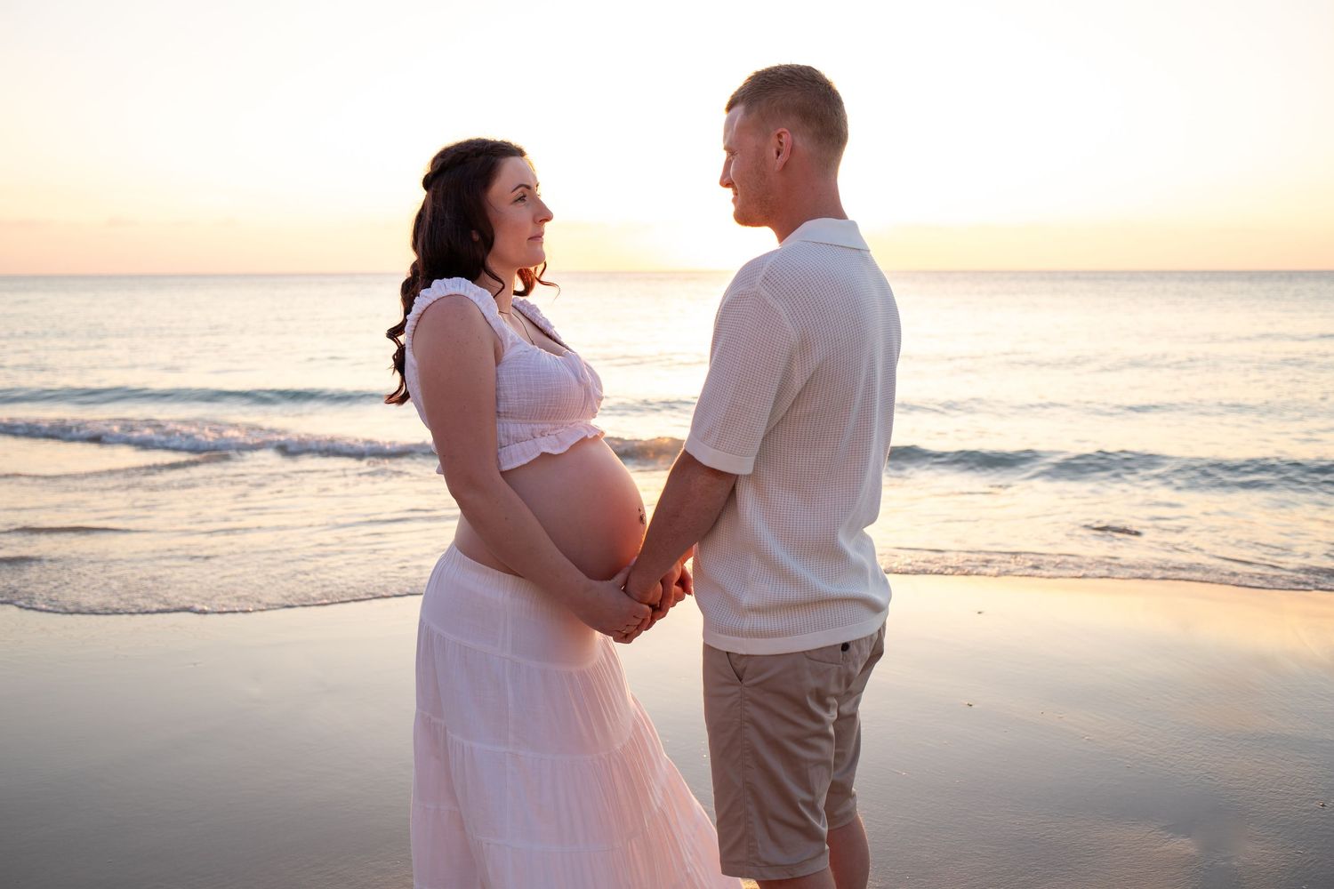 Beach maternity photoshoot at sunset with couple holding hands by the ocean.