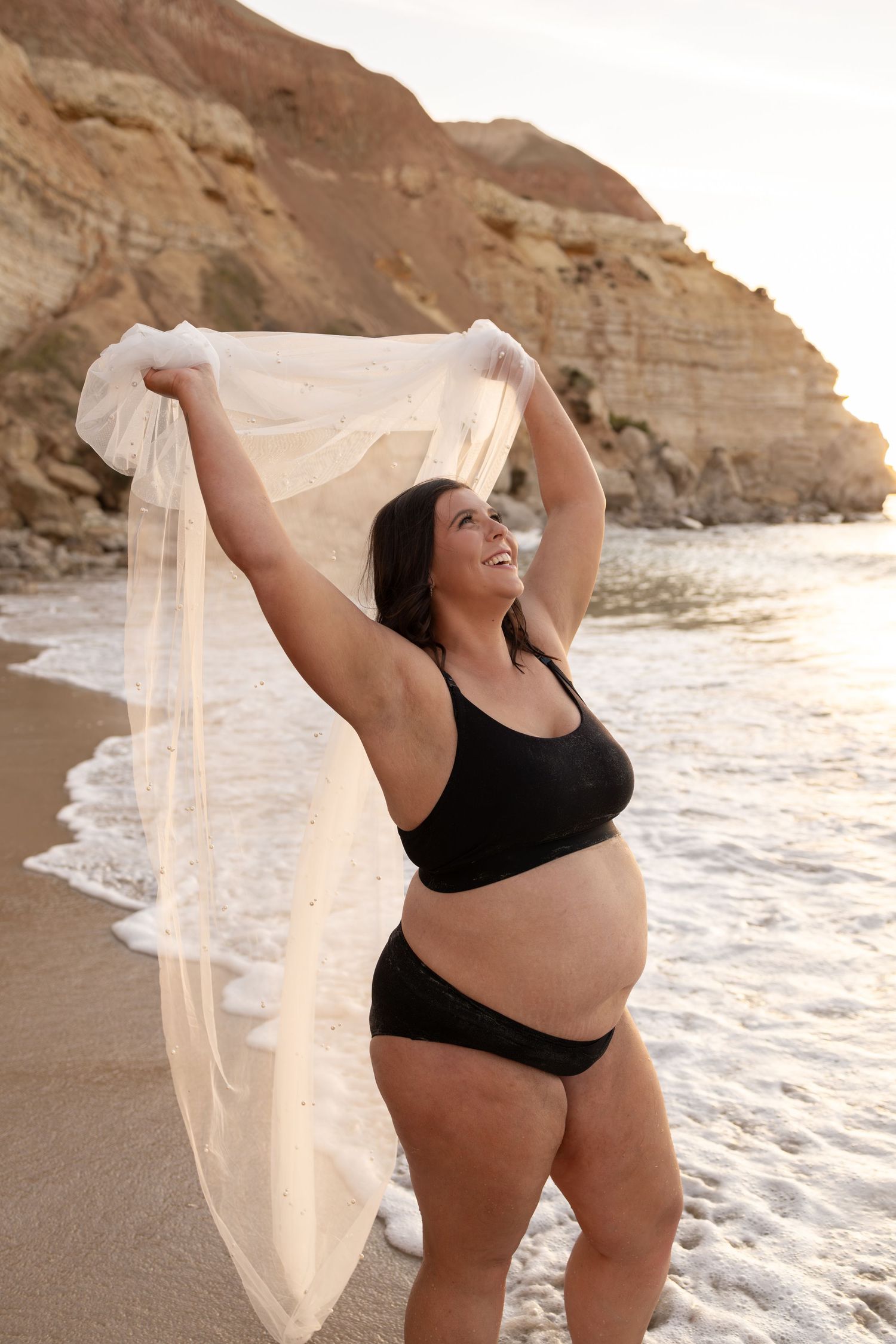 Body positive maternity beach photos with flowing fabric against ocean backdrop at sunset.