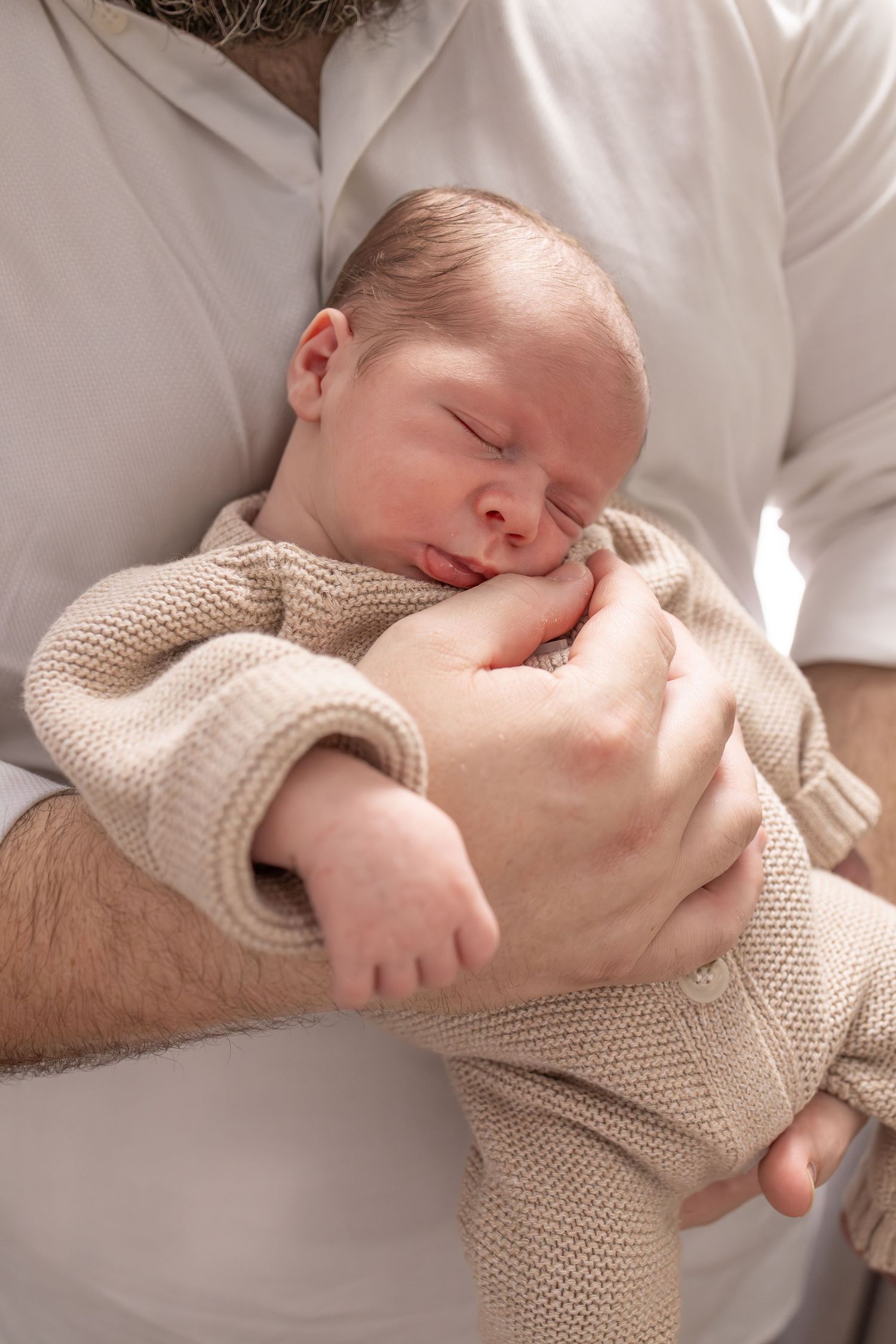 Sleeping newborn baby wearing a knit onesie held in fathers loving arms.