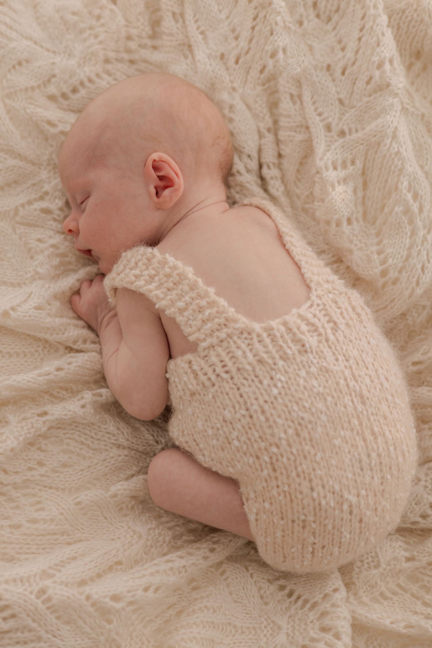 Sleeping newborn wearing knitted cream romper on textured white blanket.