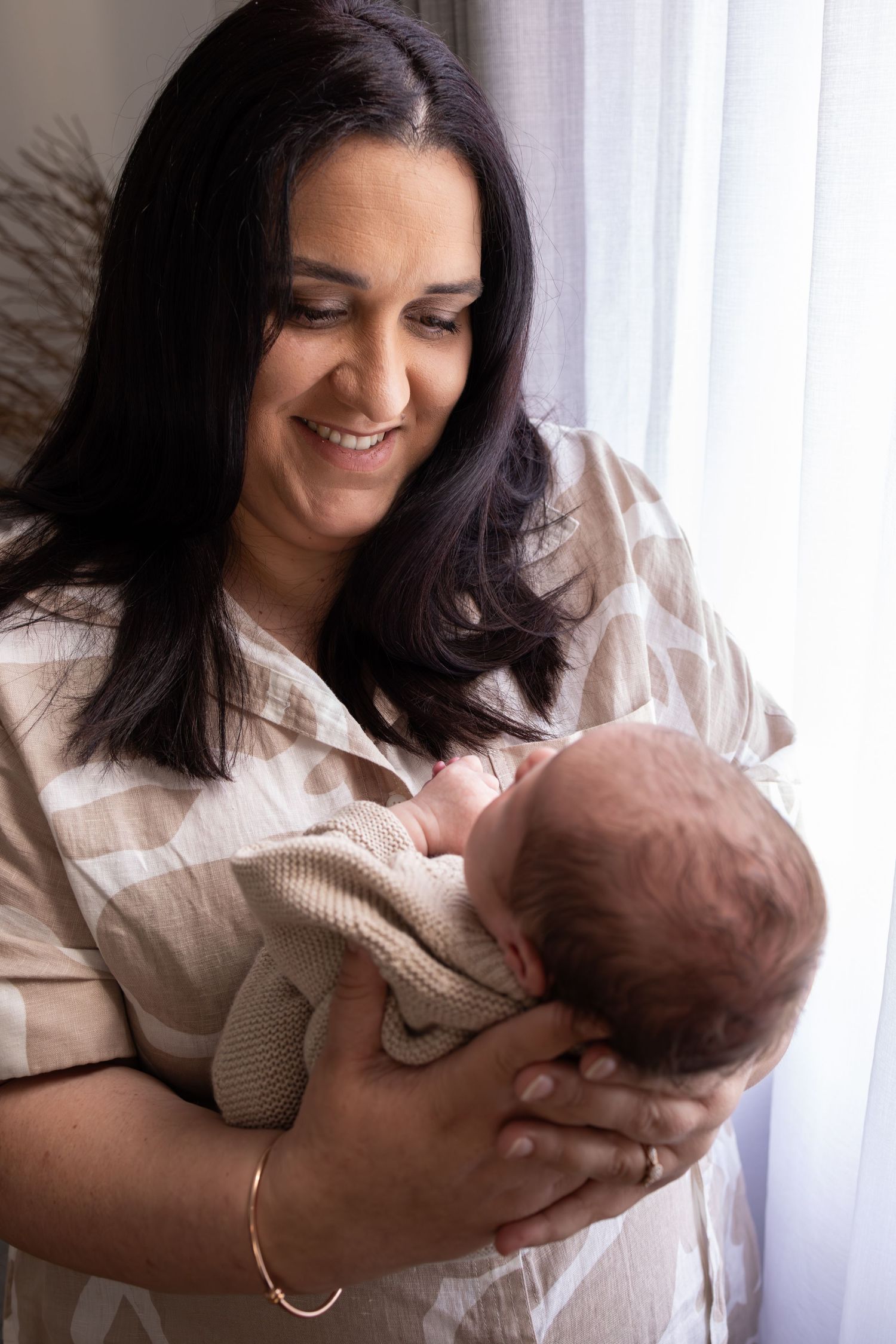 Mother in white patterned dress lovingly holds sleeping newborn near window.