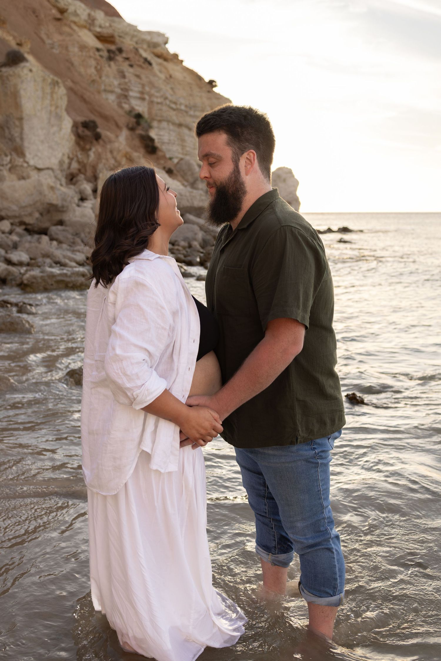 Romantic beach maternity photos with couple in white skirt and dark shirt against rocky coastline.