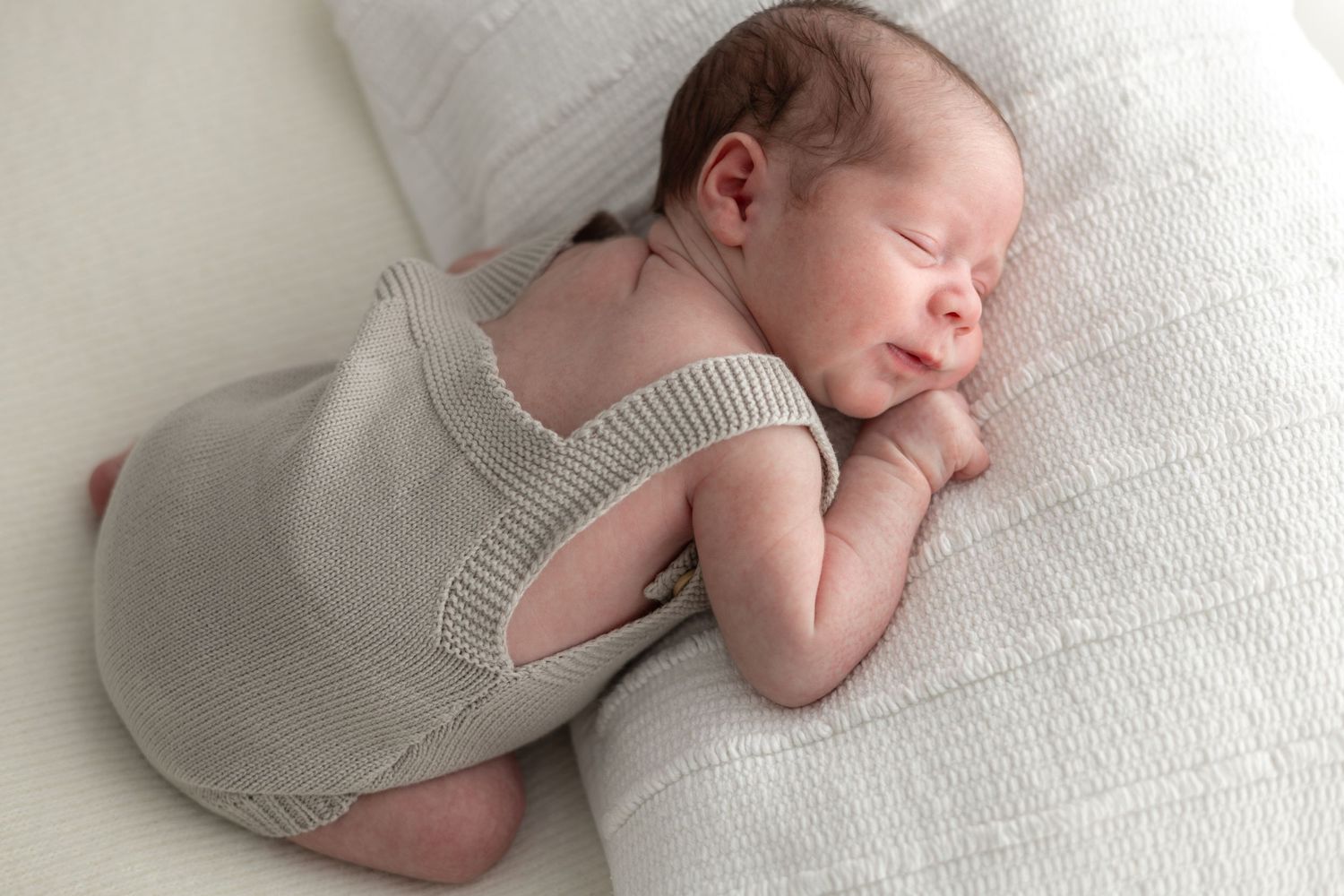 Newborn wearing knit romper resting on textured white cushion.