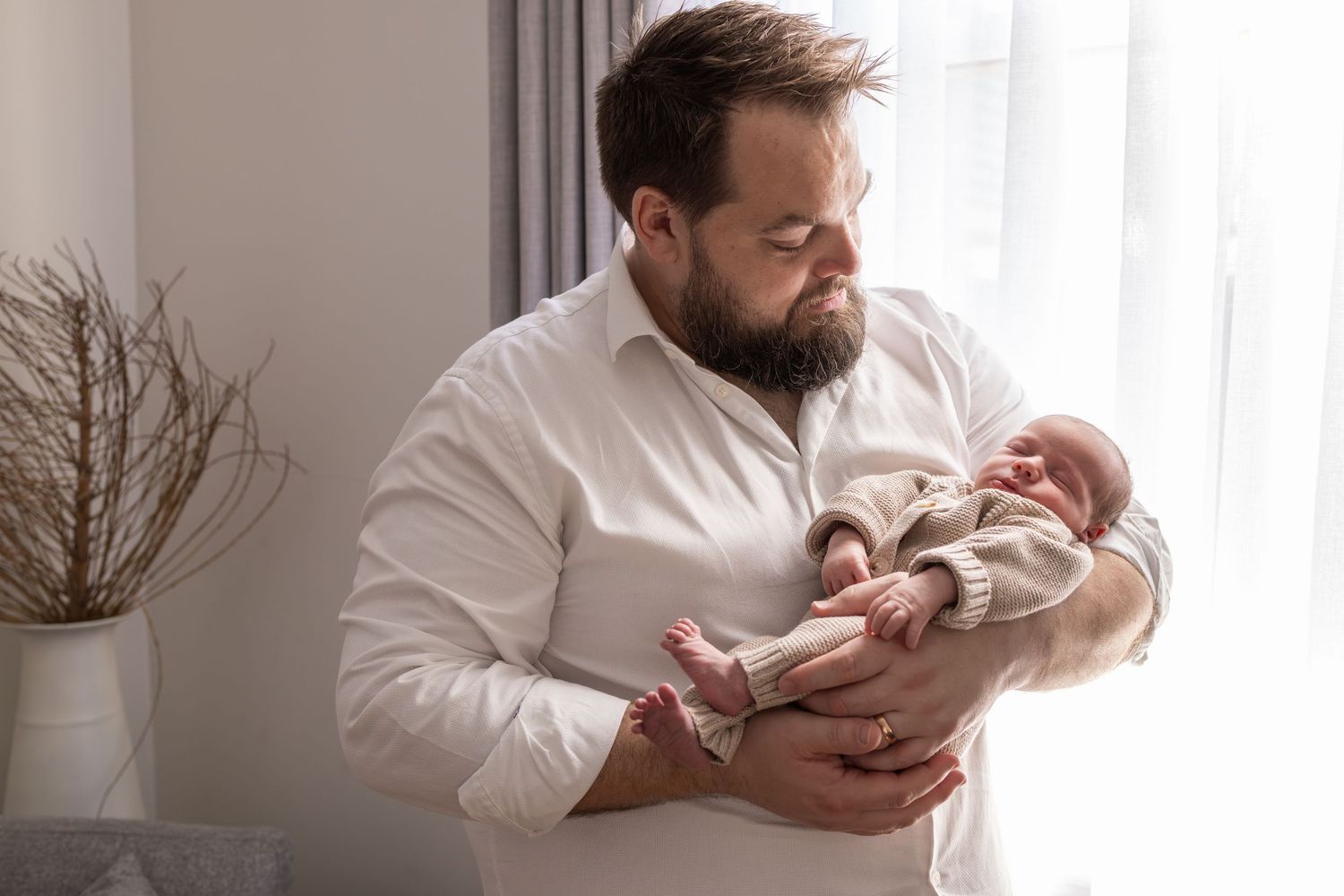 Tender moment of father cradling newborn baby in white shirt against bright window light.
