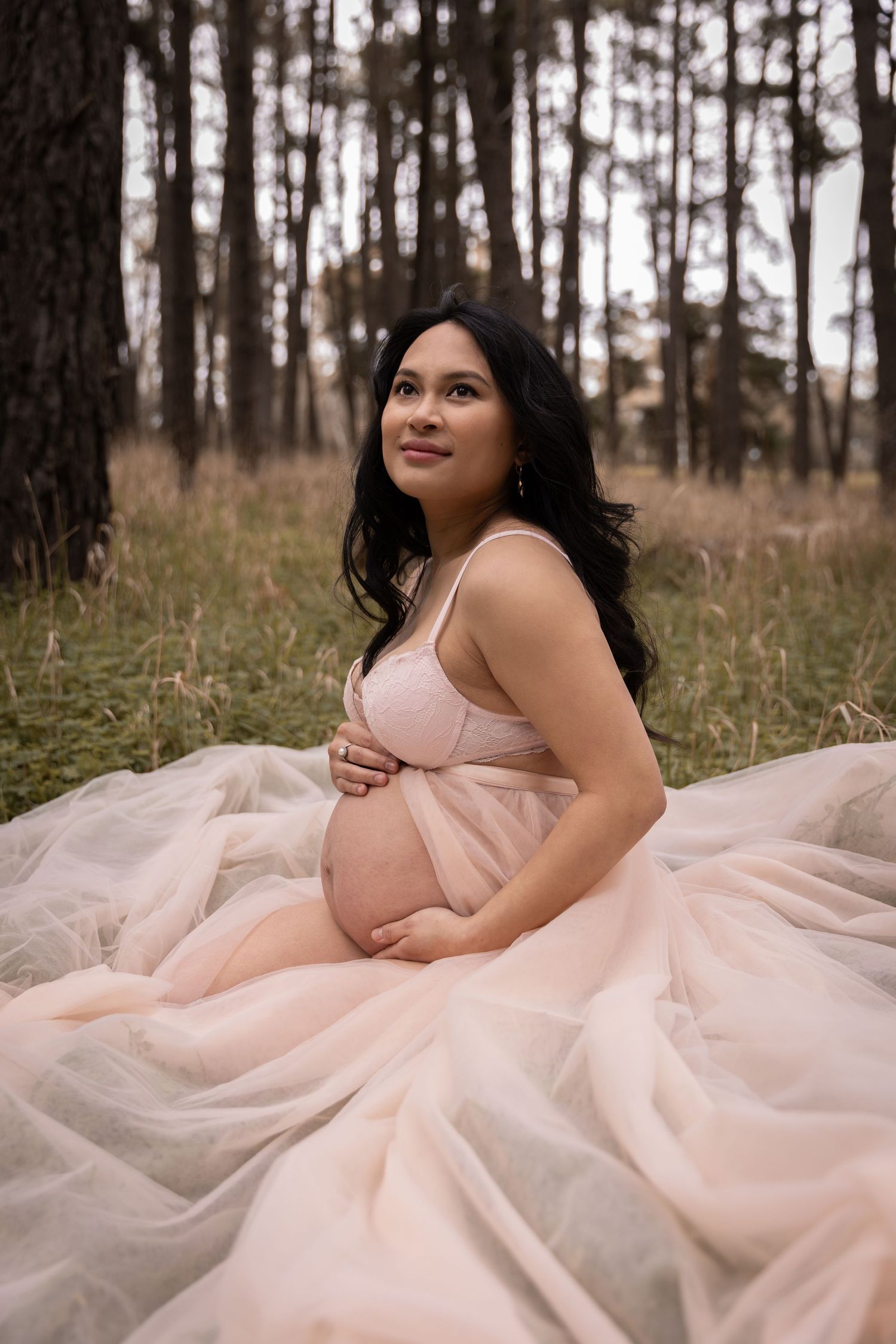 Woman in soft pink tulle gown poses gracefully in forest during a maternity photoshoot.