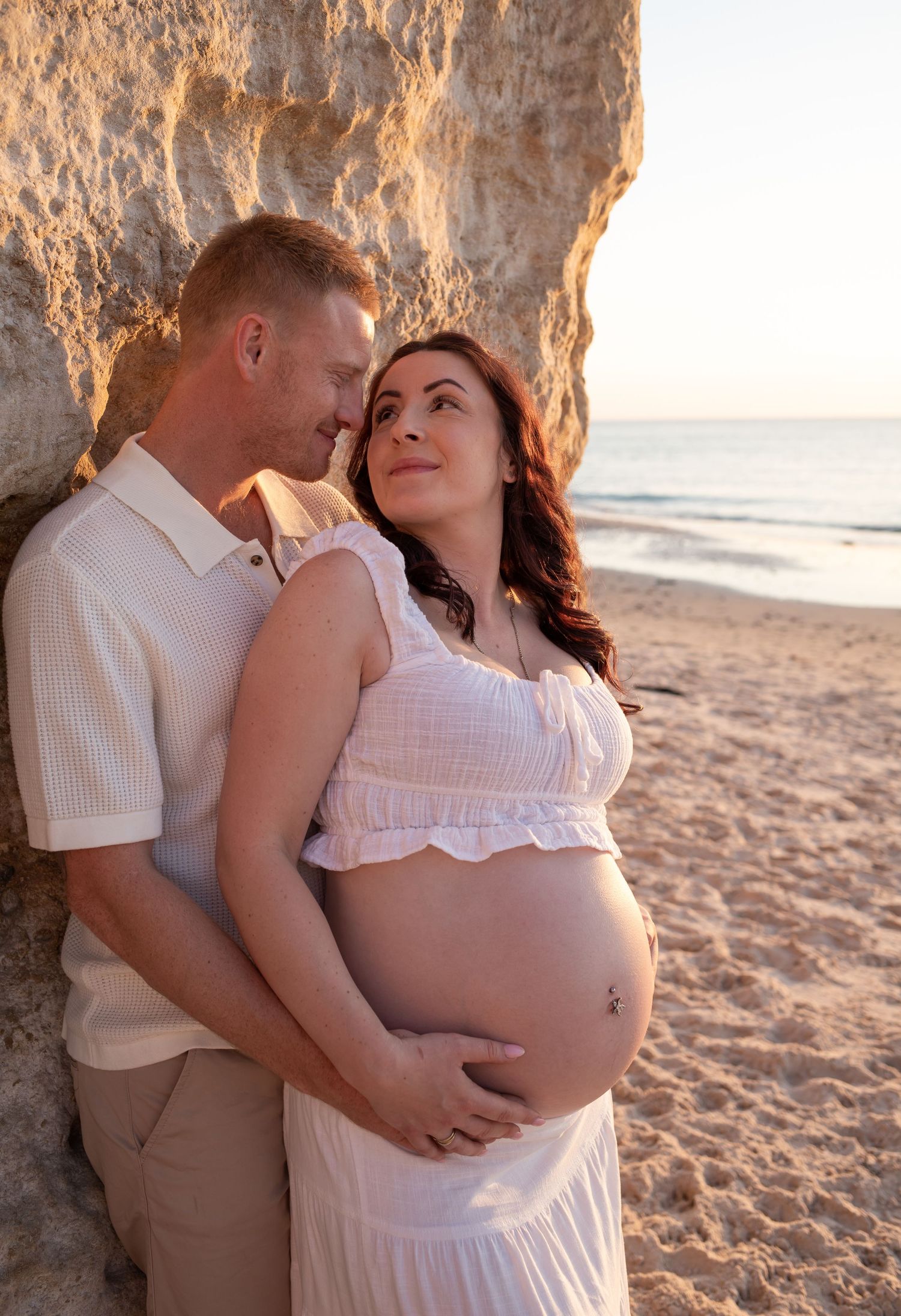 A tender maternity moment captured against a rocky beach cliff at sunset.