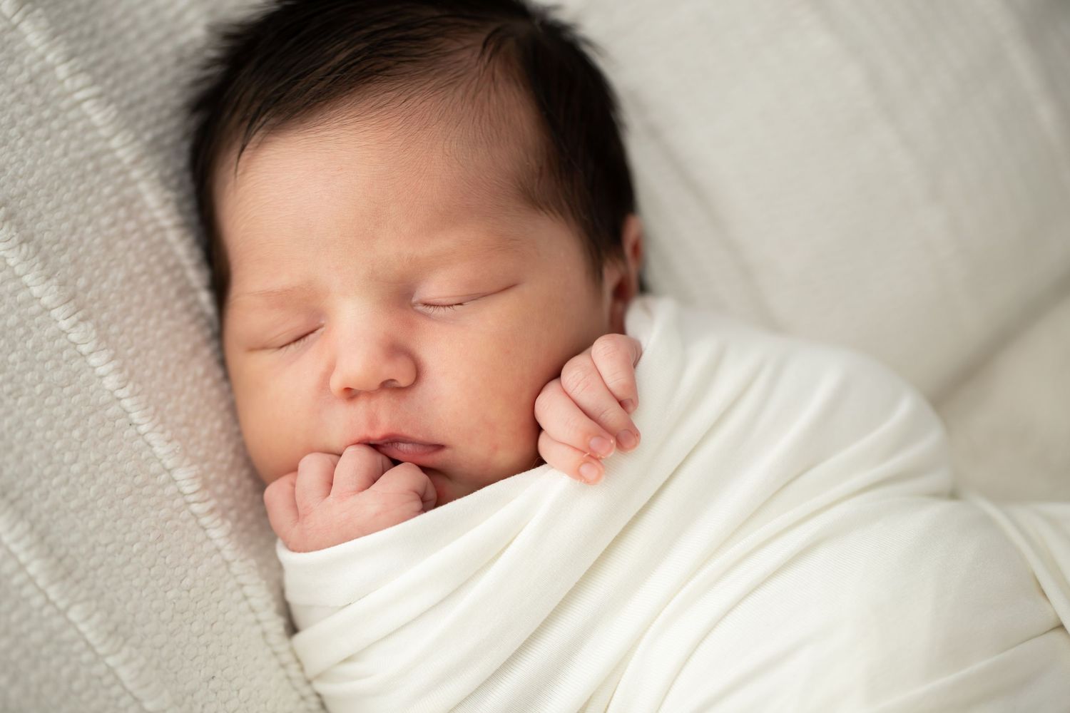 Peaceful sleeping newborn baby wrapped in white blanket with tiny hand near face.