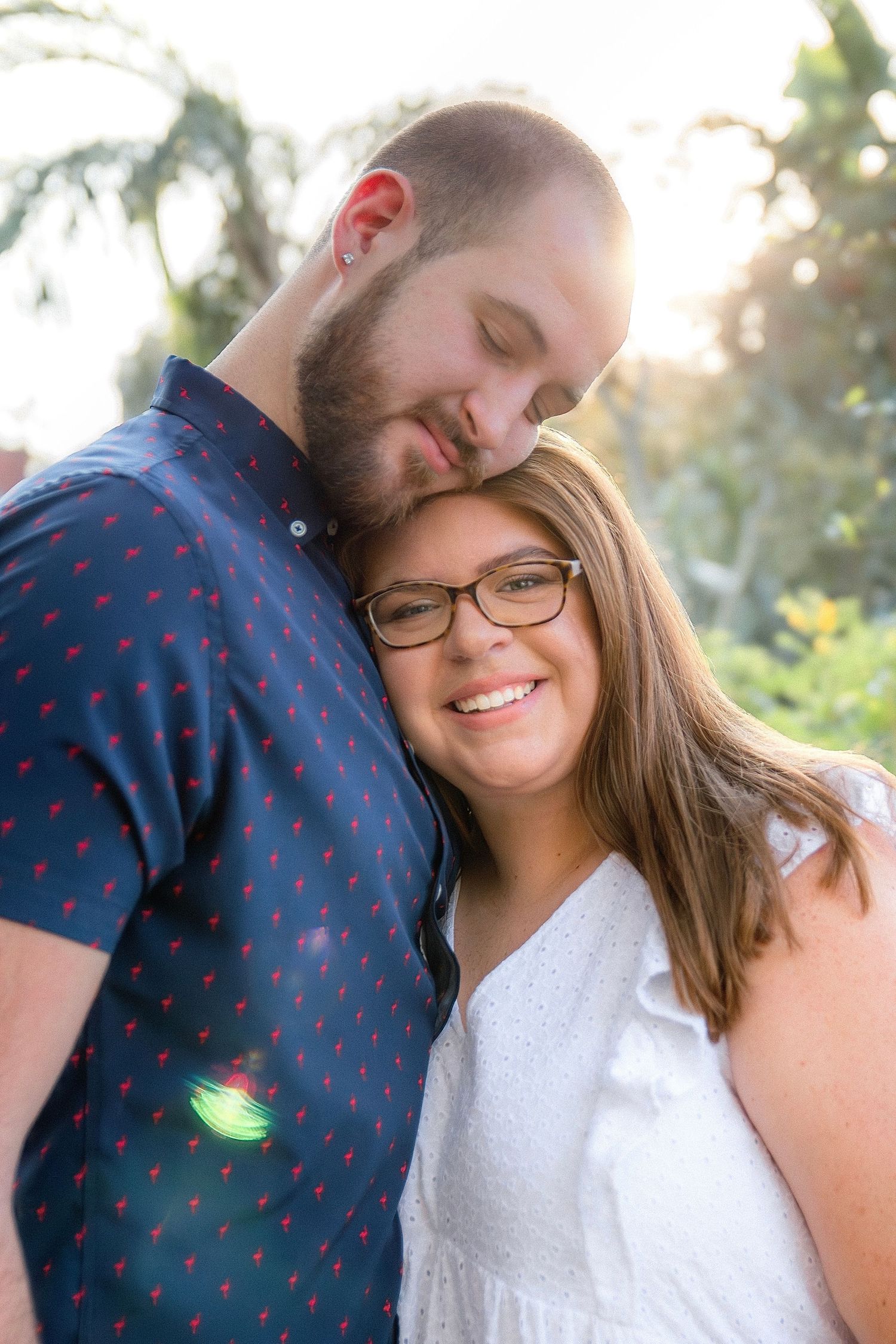 Romantic couple embraces in warm sunlight during outdoor photoshoot with natural greenery backdrop.
