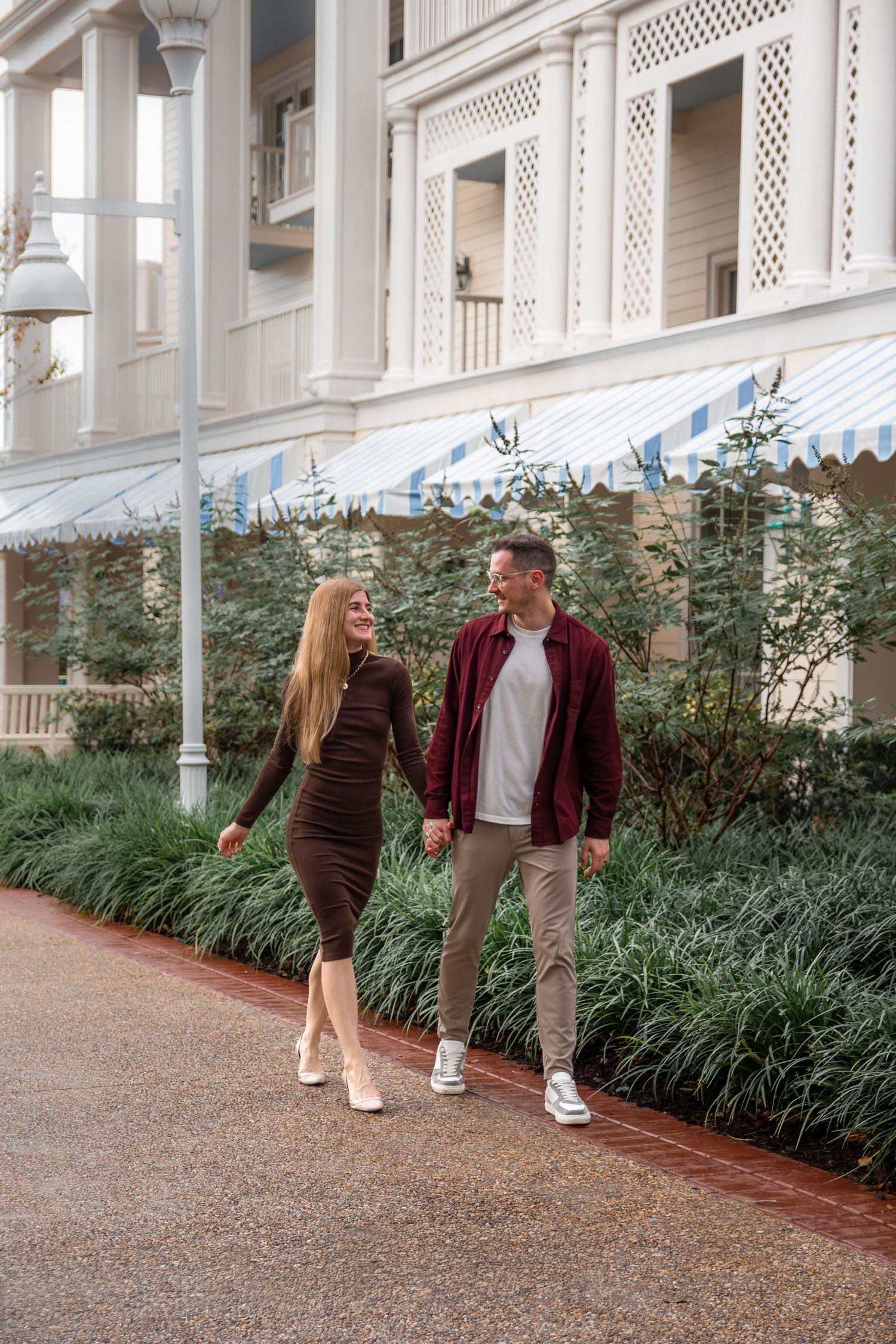 Couple walking together on path beside ornate white building with lattice details and lush garden landscaping.