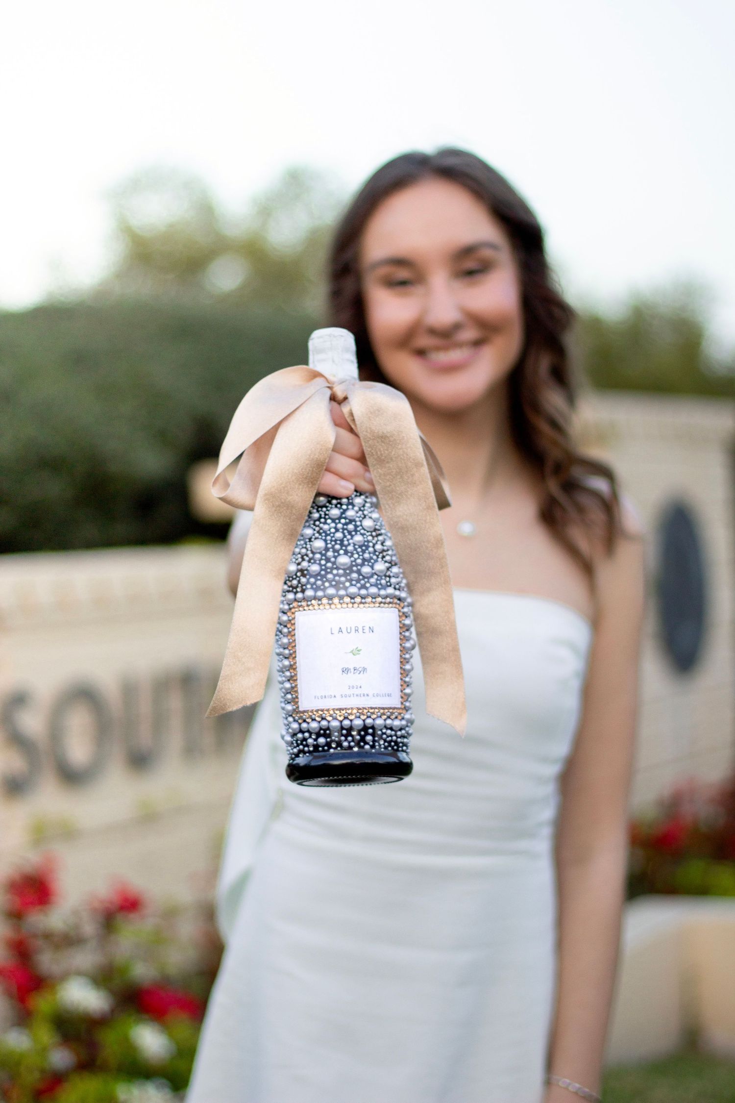 A person in a white dress holds up a small gift or favor box with a decorative bow against a garden backdrop.