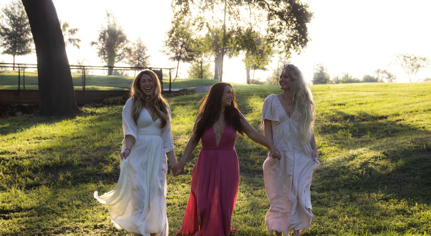 Three people in white and pink dresses walk together in a sunlit grassy field at sunset.