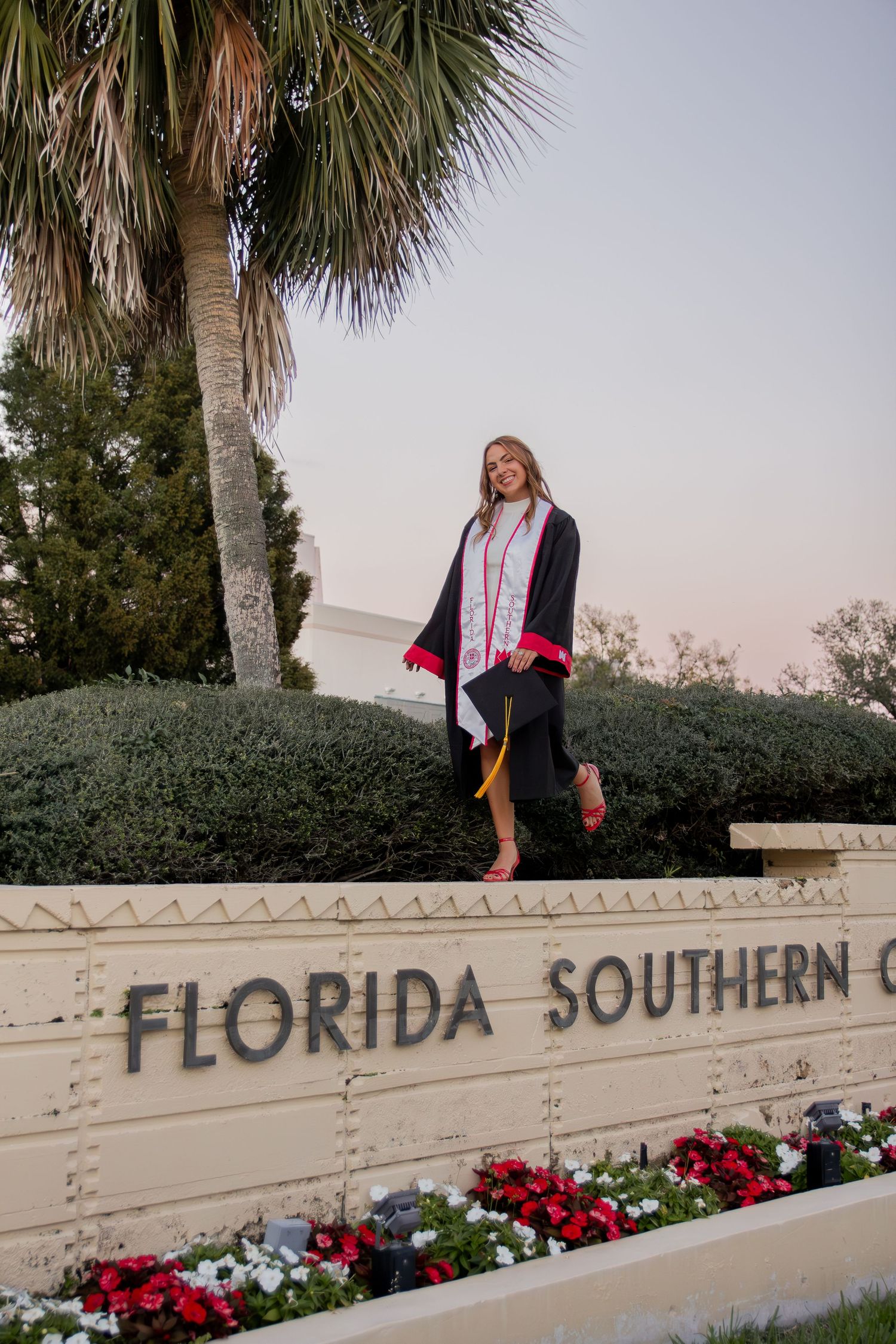 Graduate poses on Florida Southern College entrance wall decorated with vibrant red and white flowers.